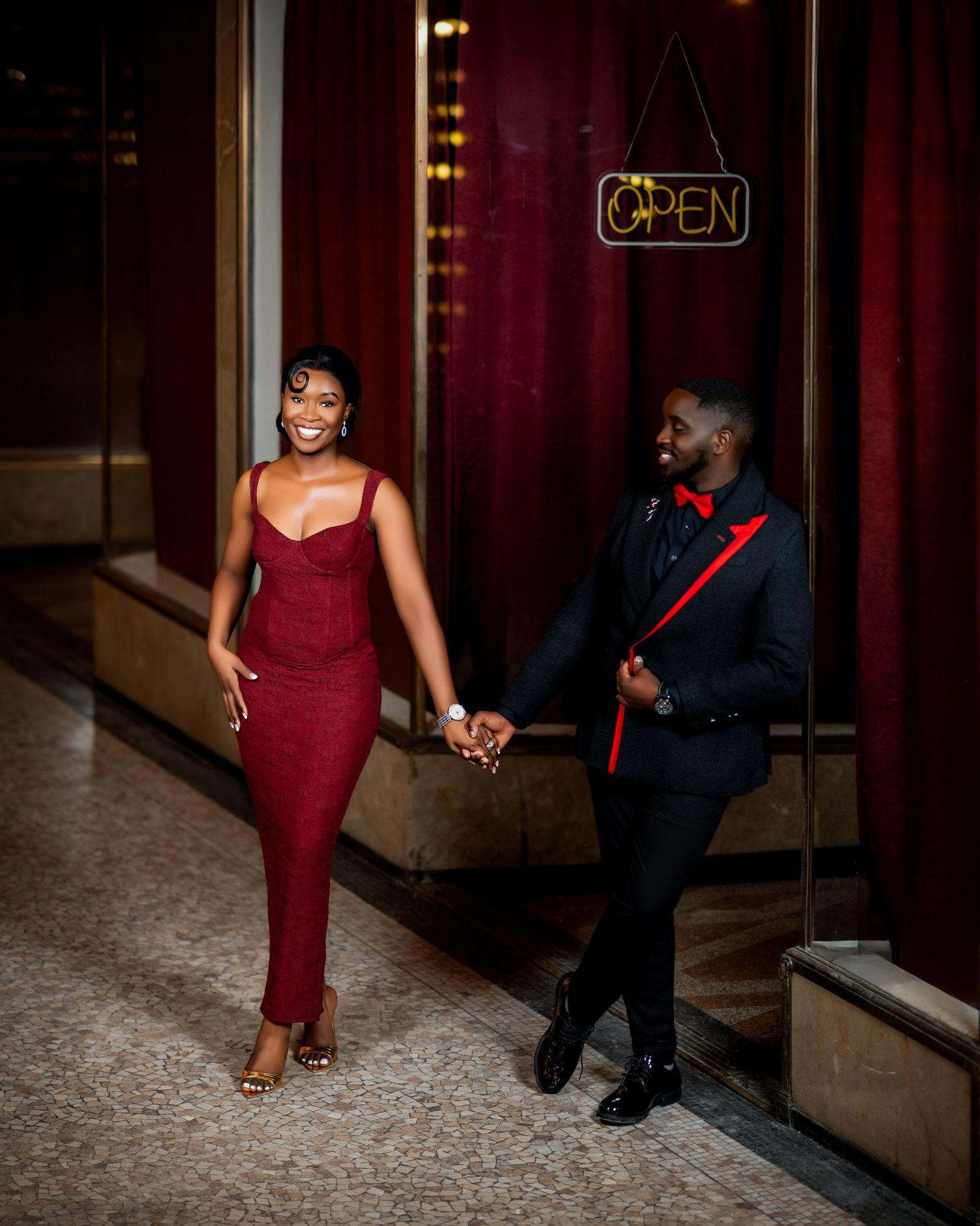 Joyful couple in burgundy dress and black suit walking hand in hand past red curtains.