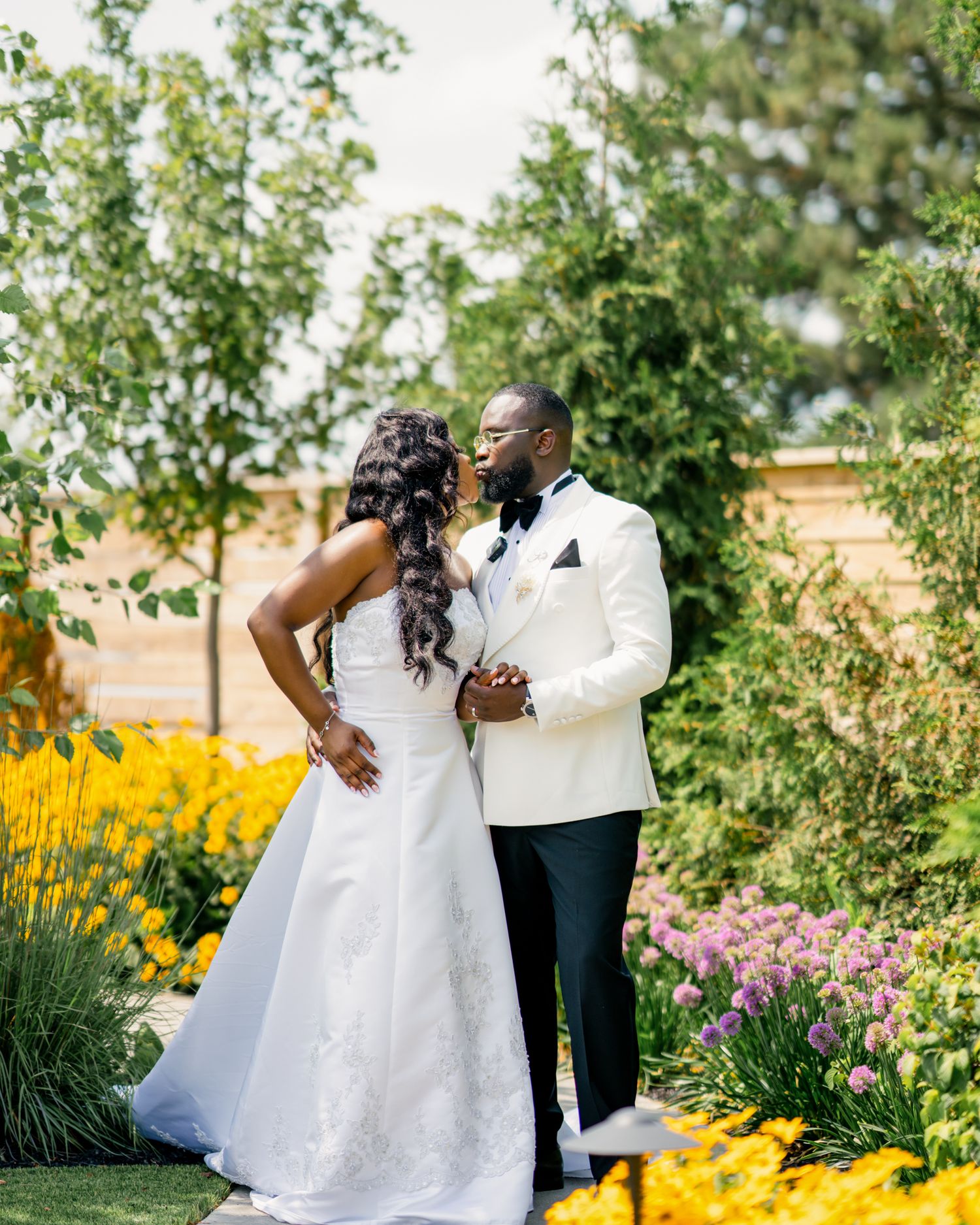 A bride and groom share a romantic moment in a lush garden, with the groom in a white tuxedo and bride in a white gown.