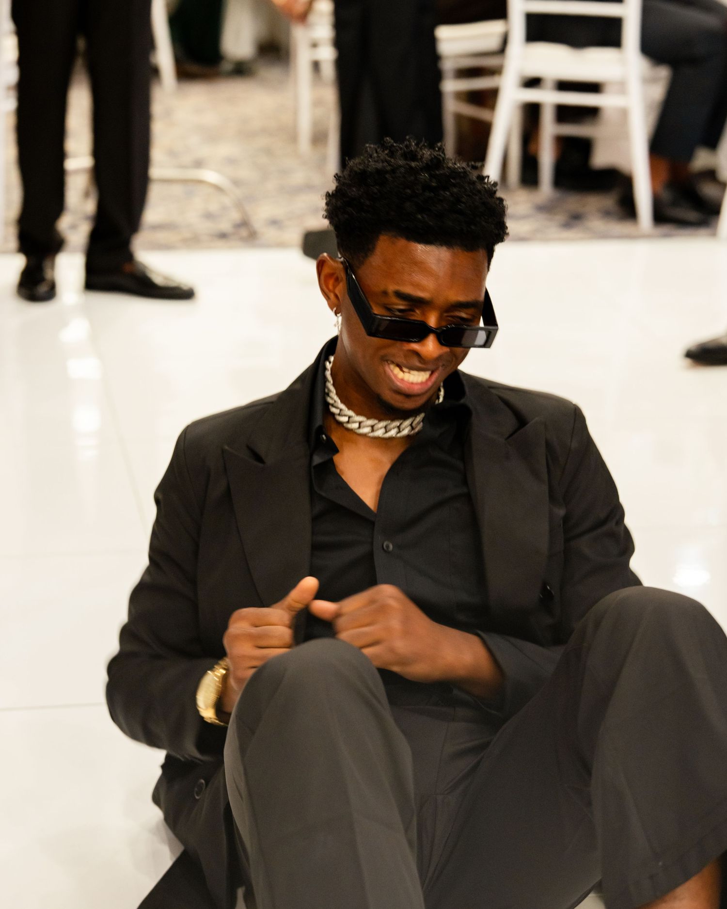 A stylish man in a black suit and gold chain necklace sits on a white floor, smiling and wearing sunglasses.