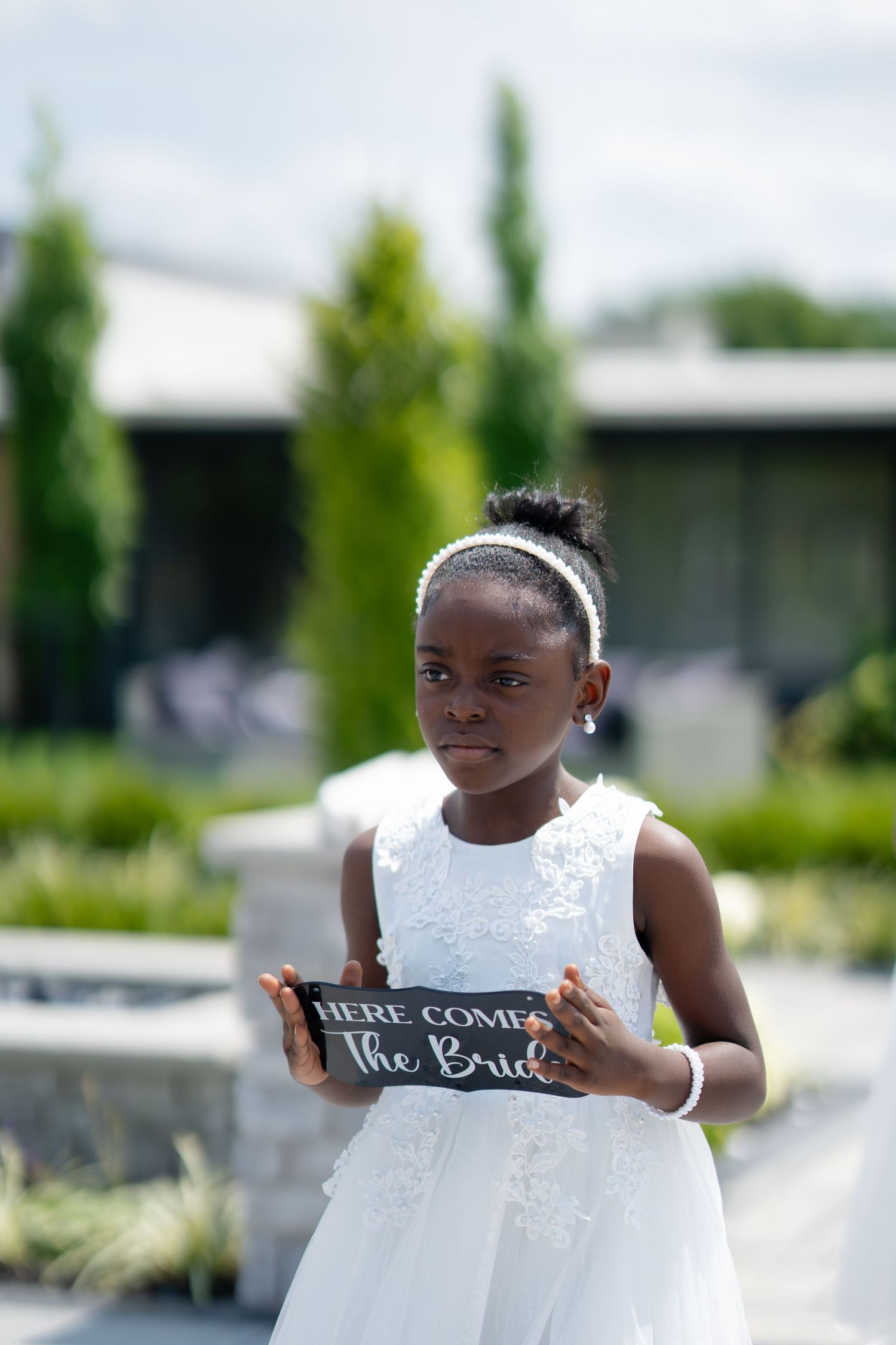 Young flower girl in white lace dress holding a Here Comes The Bride sign at outdoor wedding.