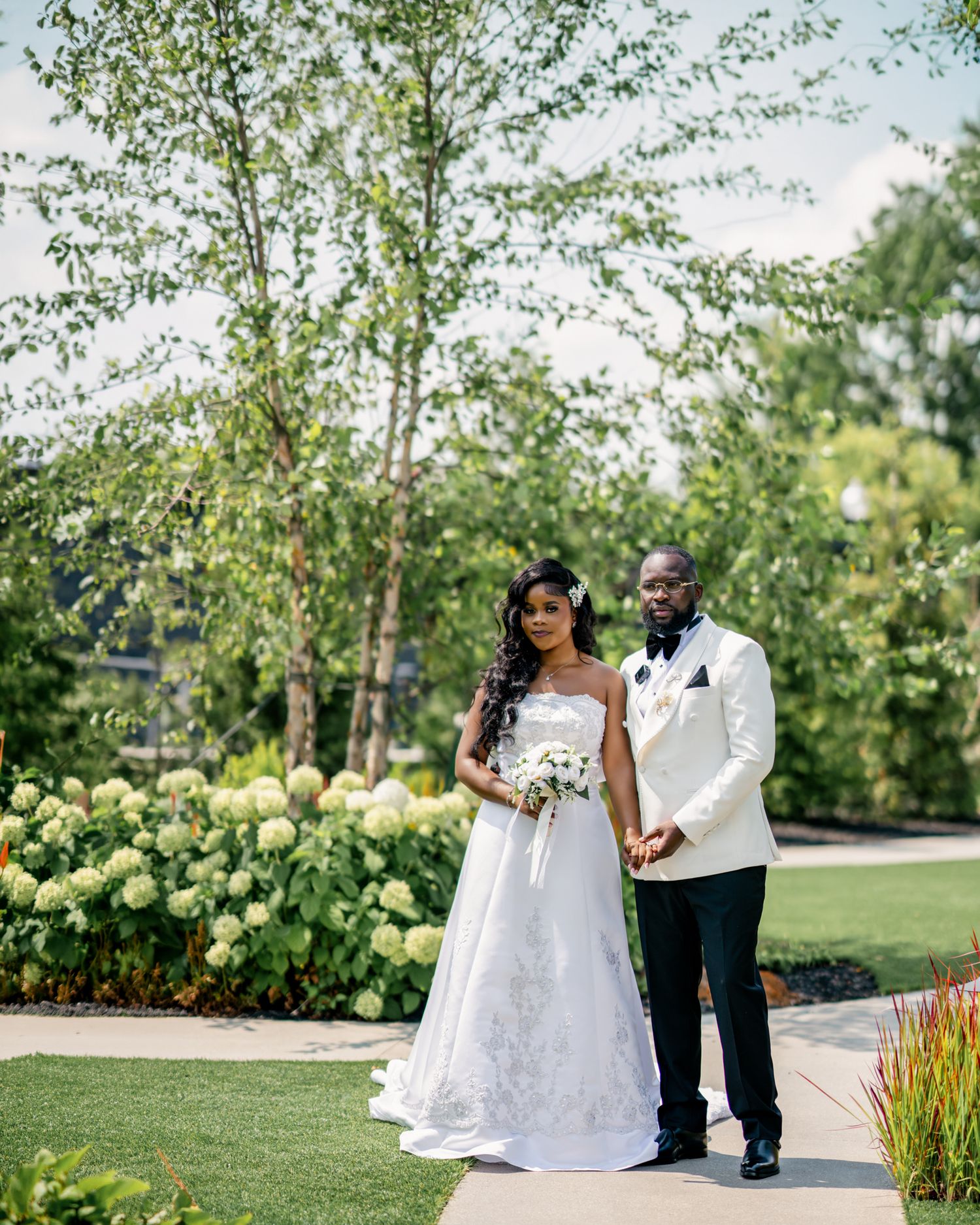 Black bride and groom in white wedding attire pose in lush garden with hydrangea blooms.