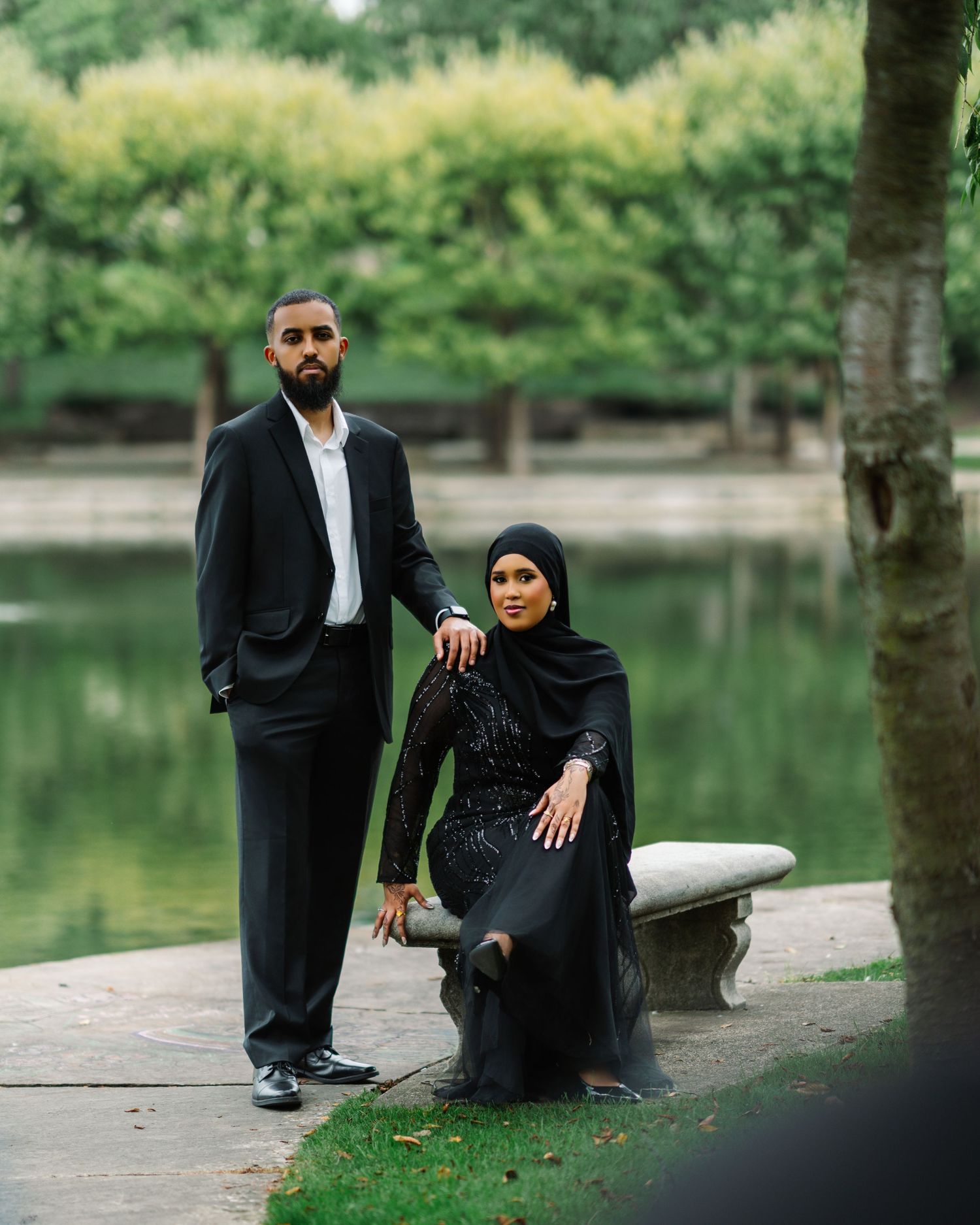 Couple in matching black formal attire posing elegantly by a serene garden pond.