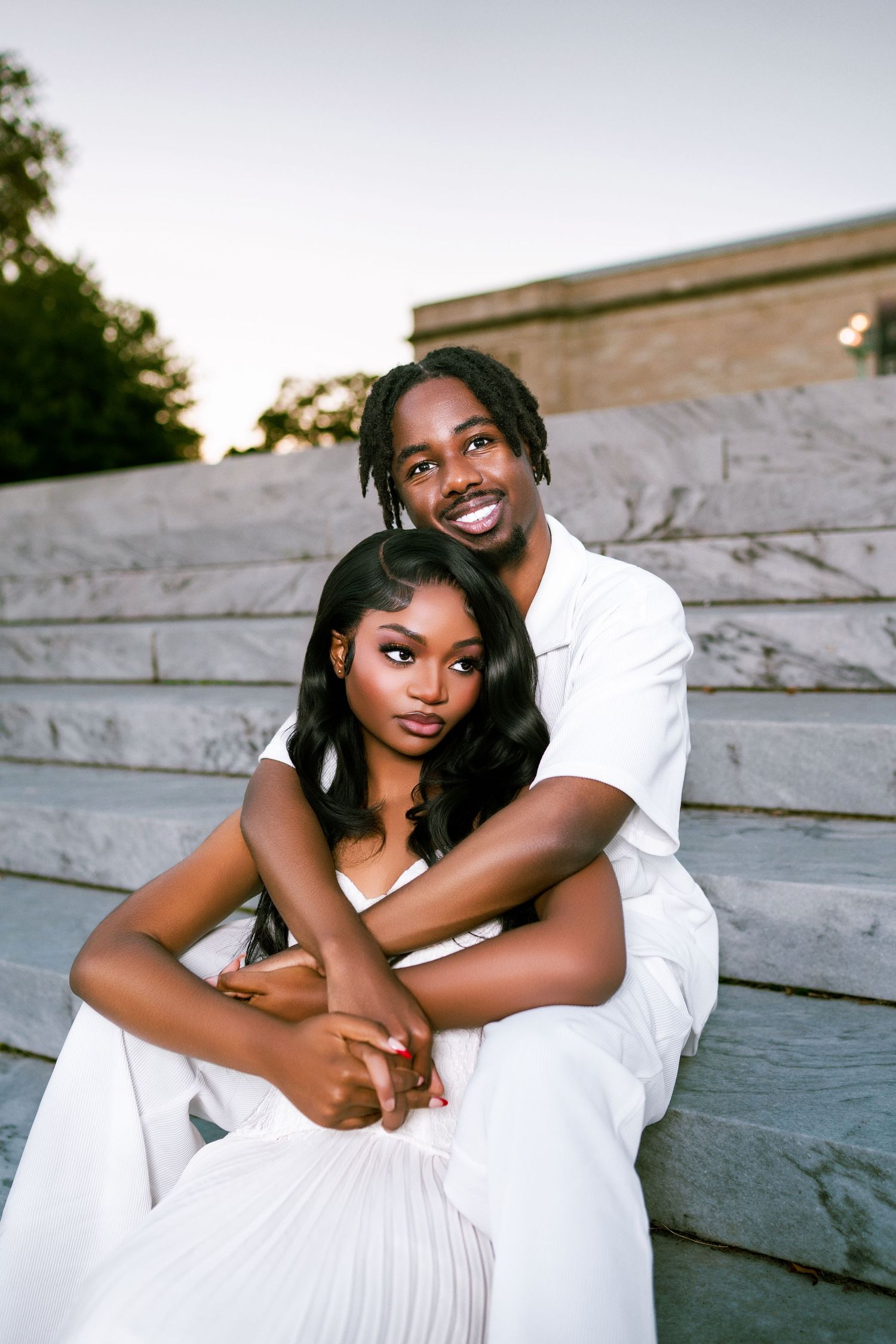 A couple in white outfits sitting on marble steps, smiling warmly during a romantic outdoor photo session.