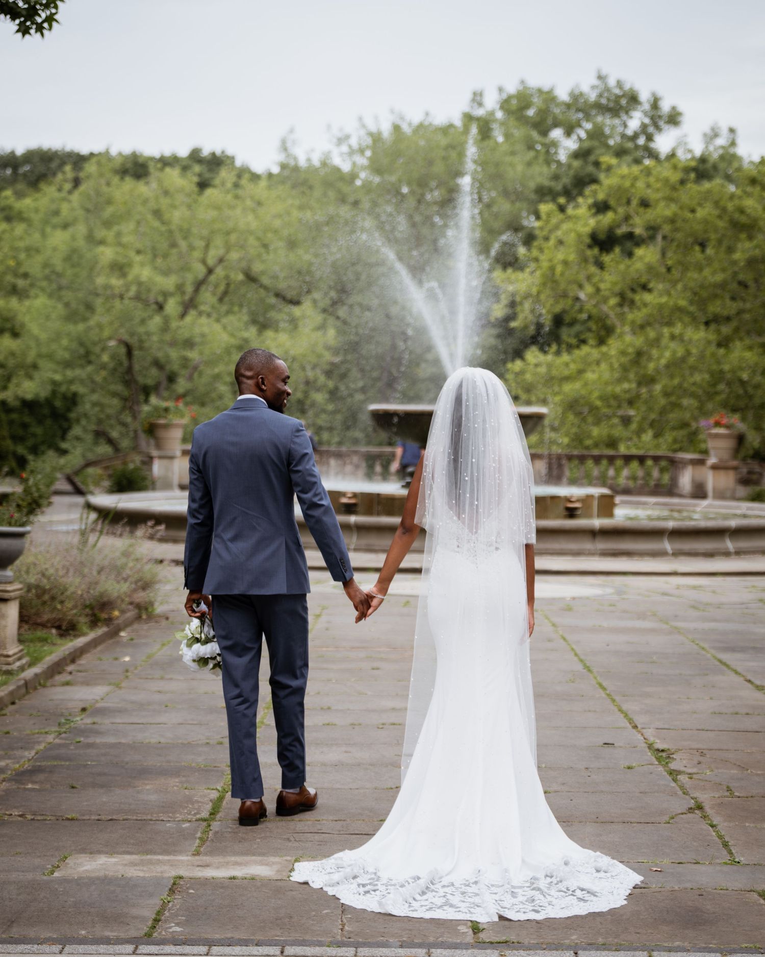 Bride and groom holding hands facing a fountain in a beautiful outdoor garden.