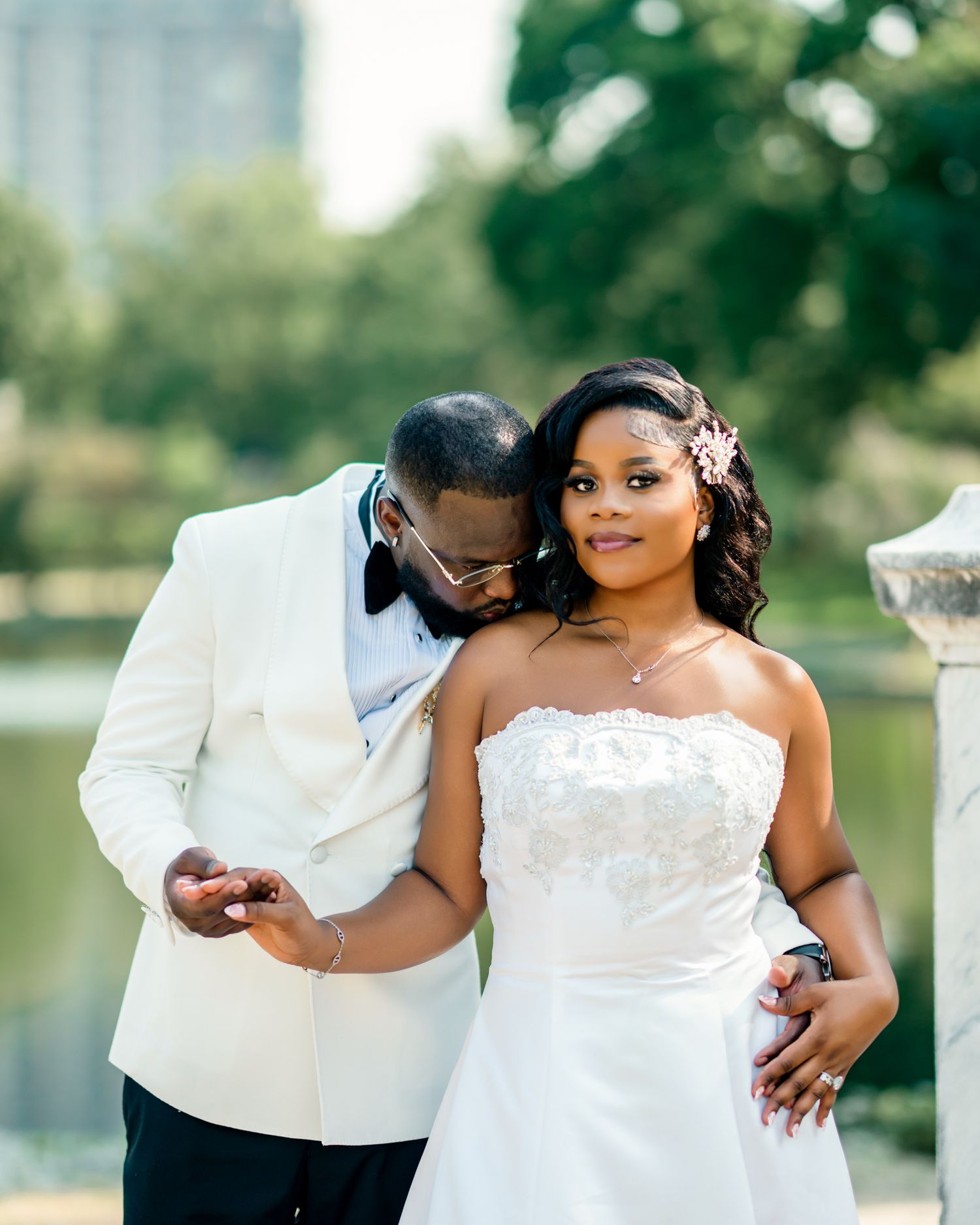 Groom in white tuxedo embraces smiling bride in strapless lace gown near a serene pond.