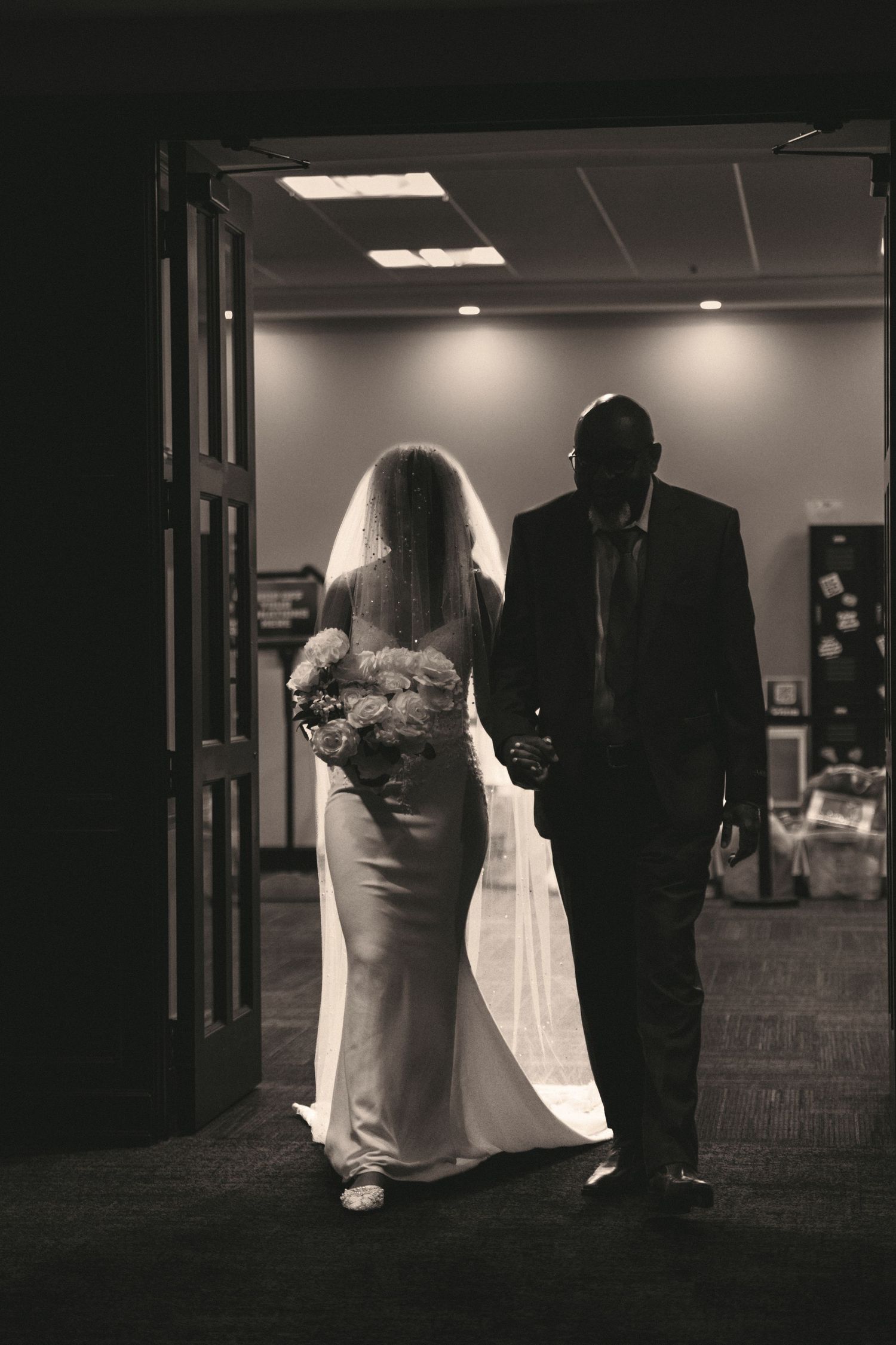 Silhouette of bride in fitted gown holding bouquet walking with escort through doorway before wedding.