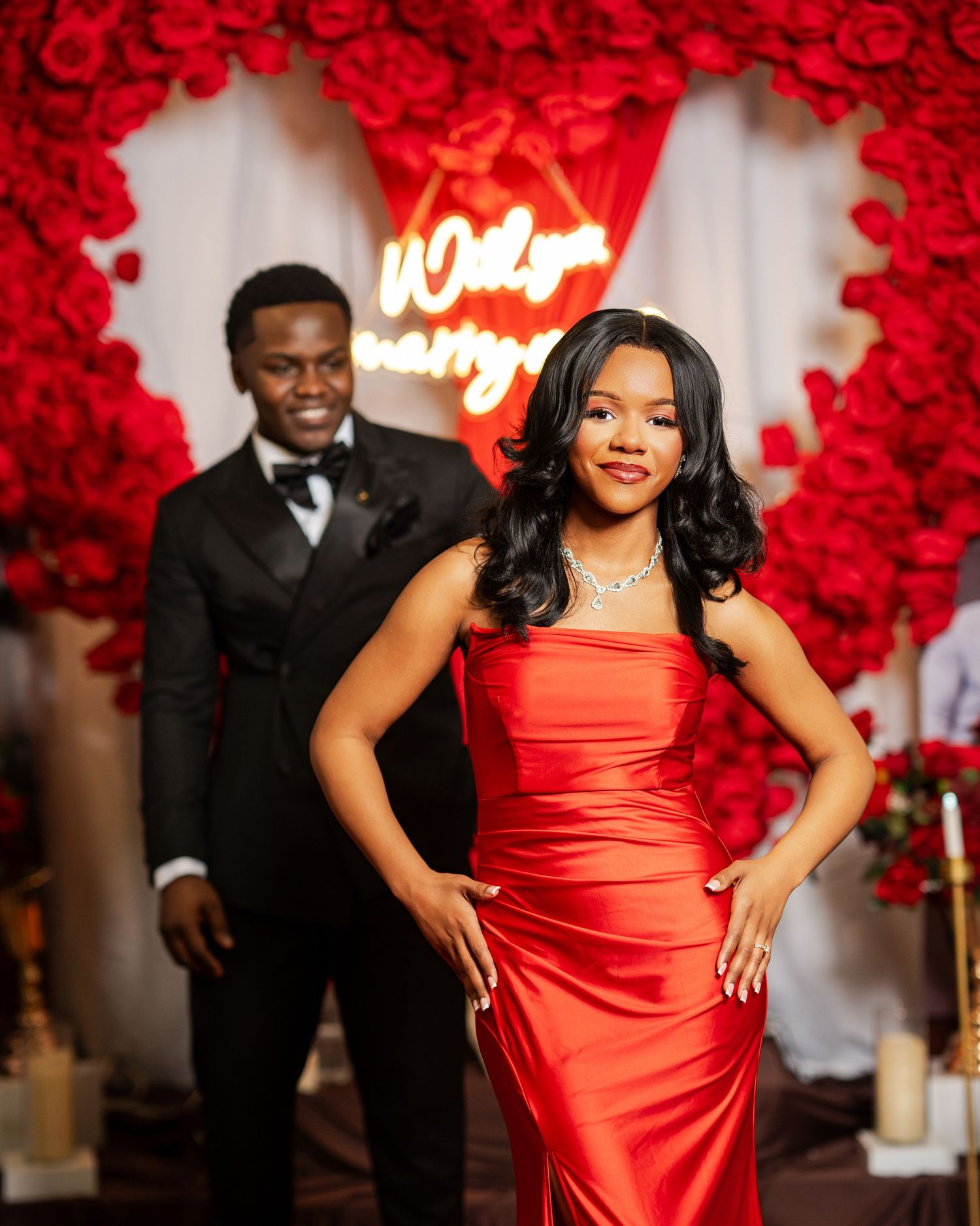 Woman in red satin gown poses at romantic proposal setup with red roses and Will You Marry Me neon sign.