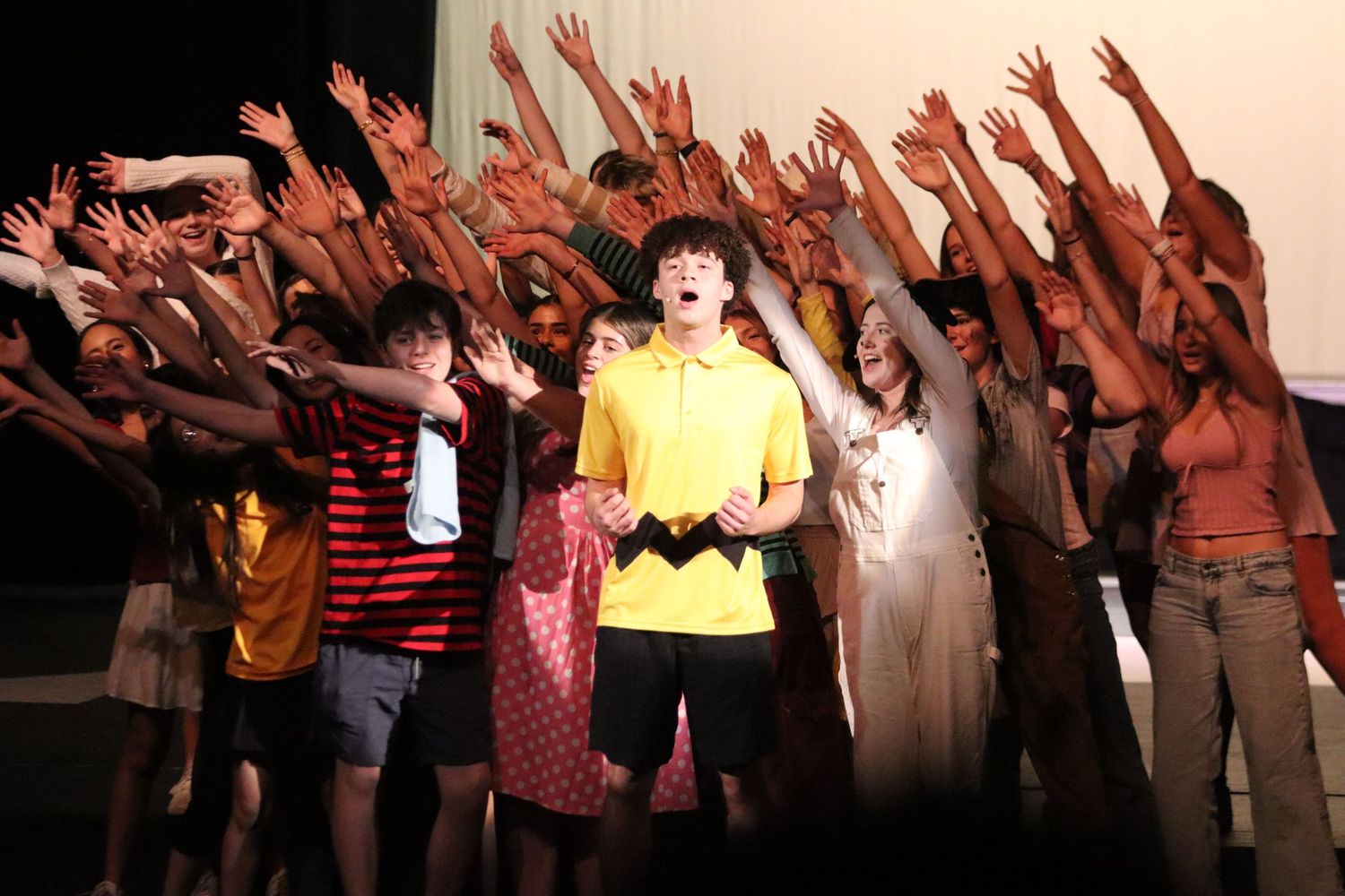 Students perform energetic musical theater production with raised hands on stage.