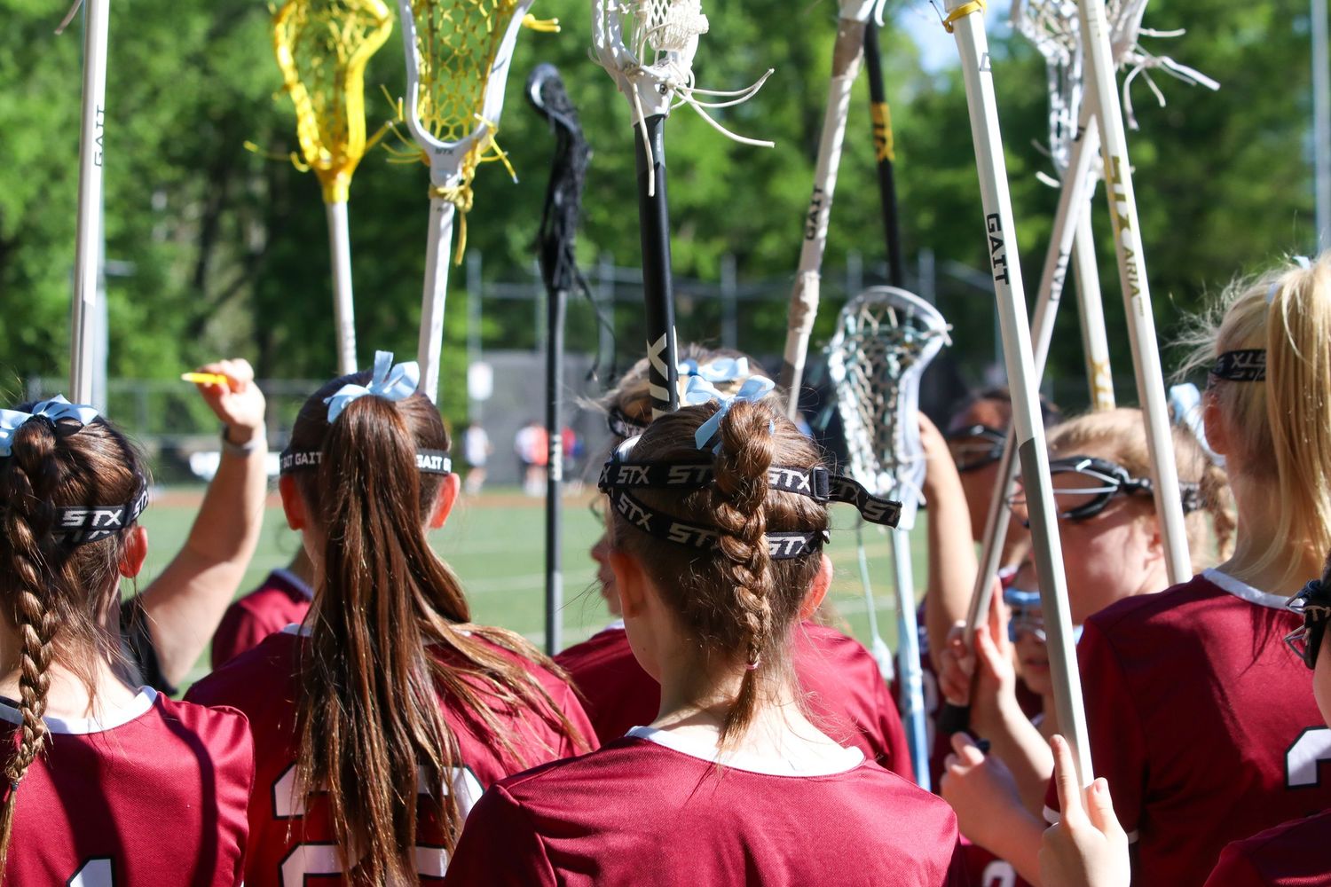 Girls lacrosse team huddles together raising sticks in maroon uniforms on sunny field.