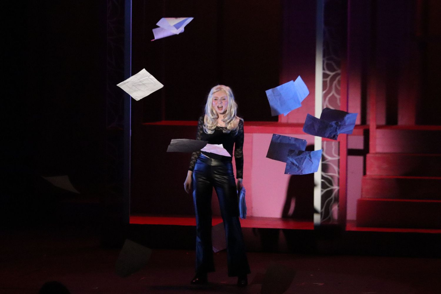Blonde performer on stage surrounded by flying papers during dramatic theatrical performance.