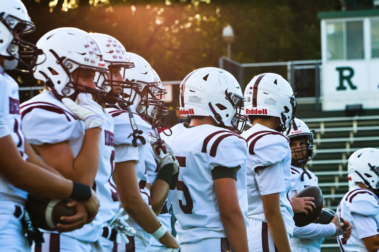 Youth football players in maroon helmets and uniforms line up on the sideline.