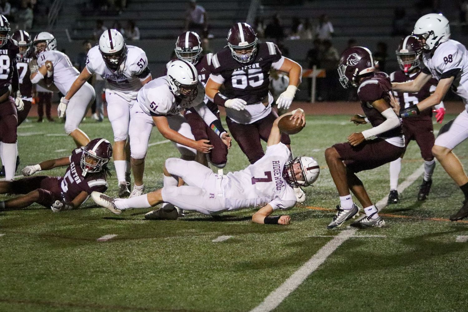 High school football players in maroon and white uniforms compete in an intense night game tackle play.