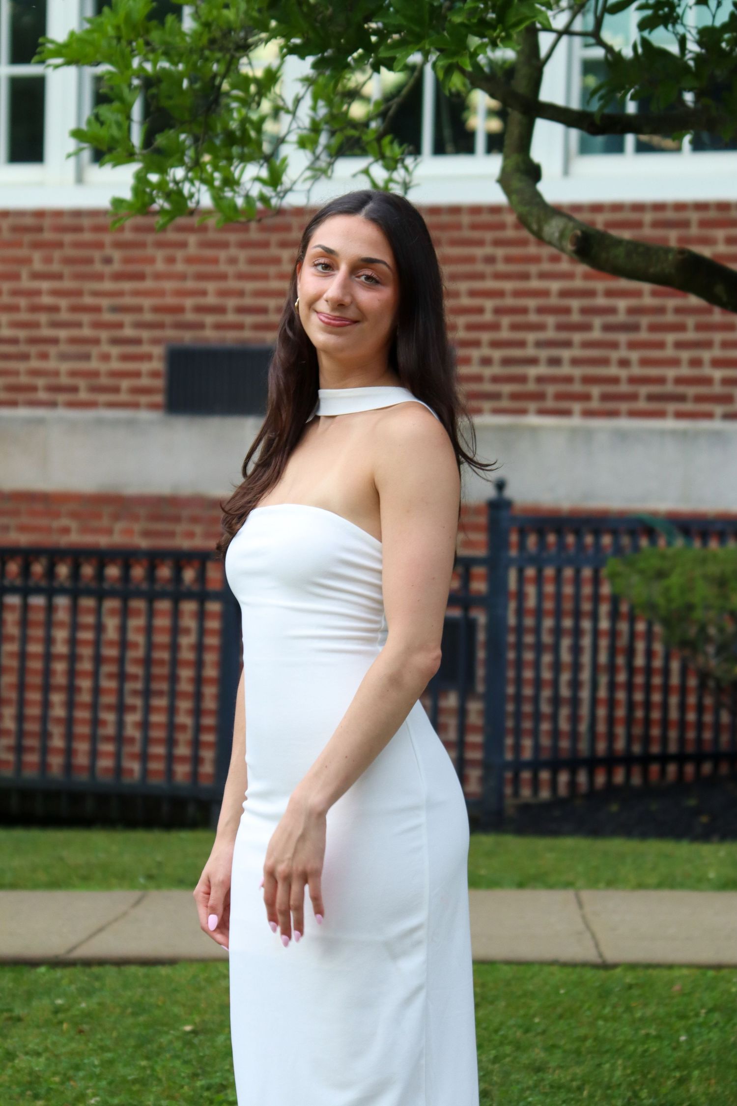 Young woman in elegant white halter dress posing outdoors near brick building and tree.