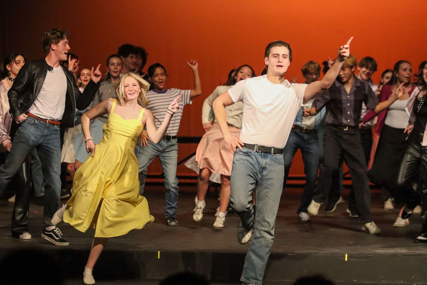 Students in 1950s costumes dance energetically in Grease school musical performance.