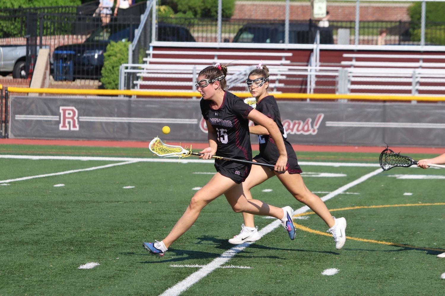 Female lacrosse player in maroon uniform sprints with the ball during a sunny game.