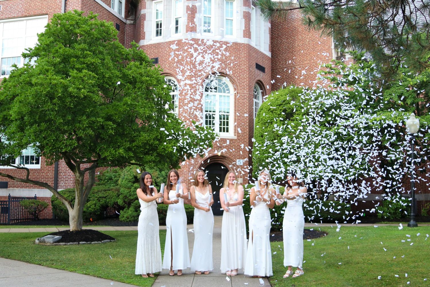 Women in white dresses celebrating with confetti explosion outside brick building.