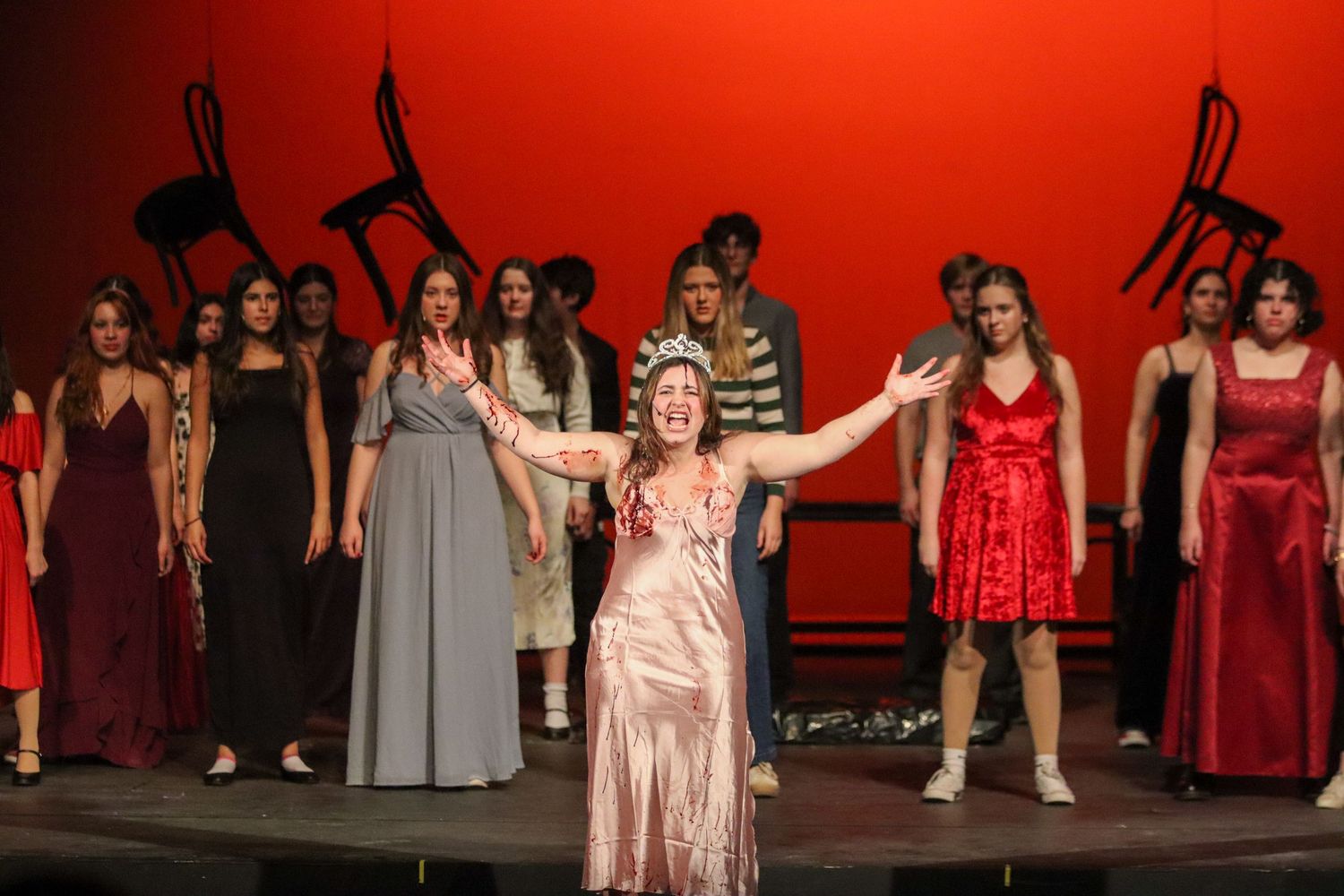 Students perform dramatic stage production in red and gray costumes under red stage lighting.