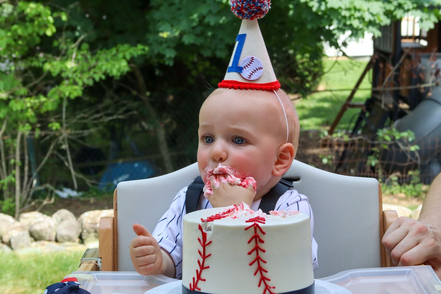 Baby in baseball-themed outfit eating first birthday cake with party hat outdoors.