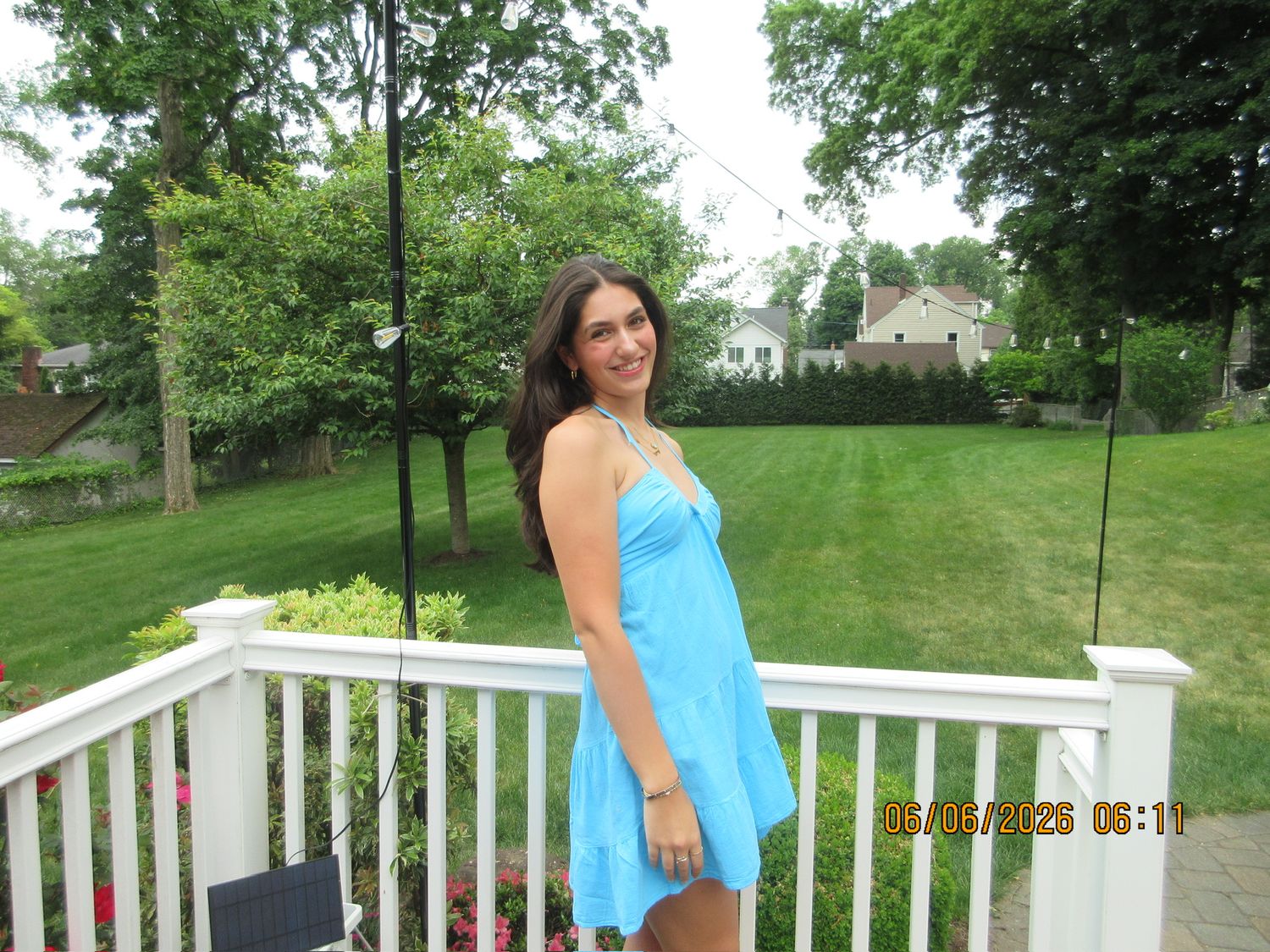 Young woman in turquoise sundress smiling on white deck surrounded by green backyard.
