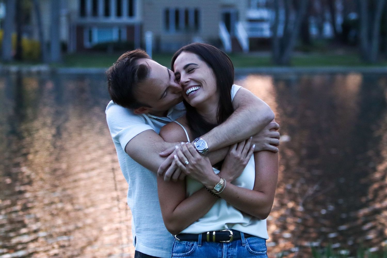 Happy couple embracing and laughing together near reflective lake at golden hour sunset.