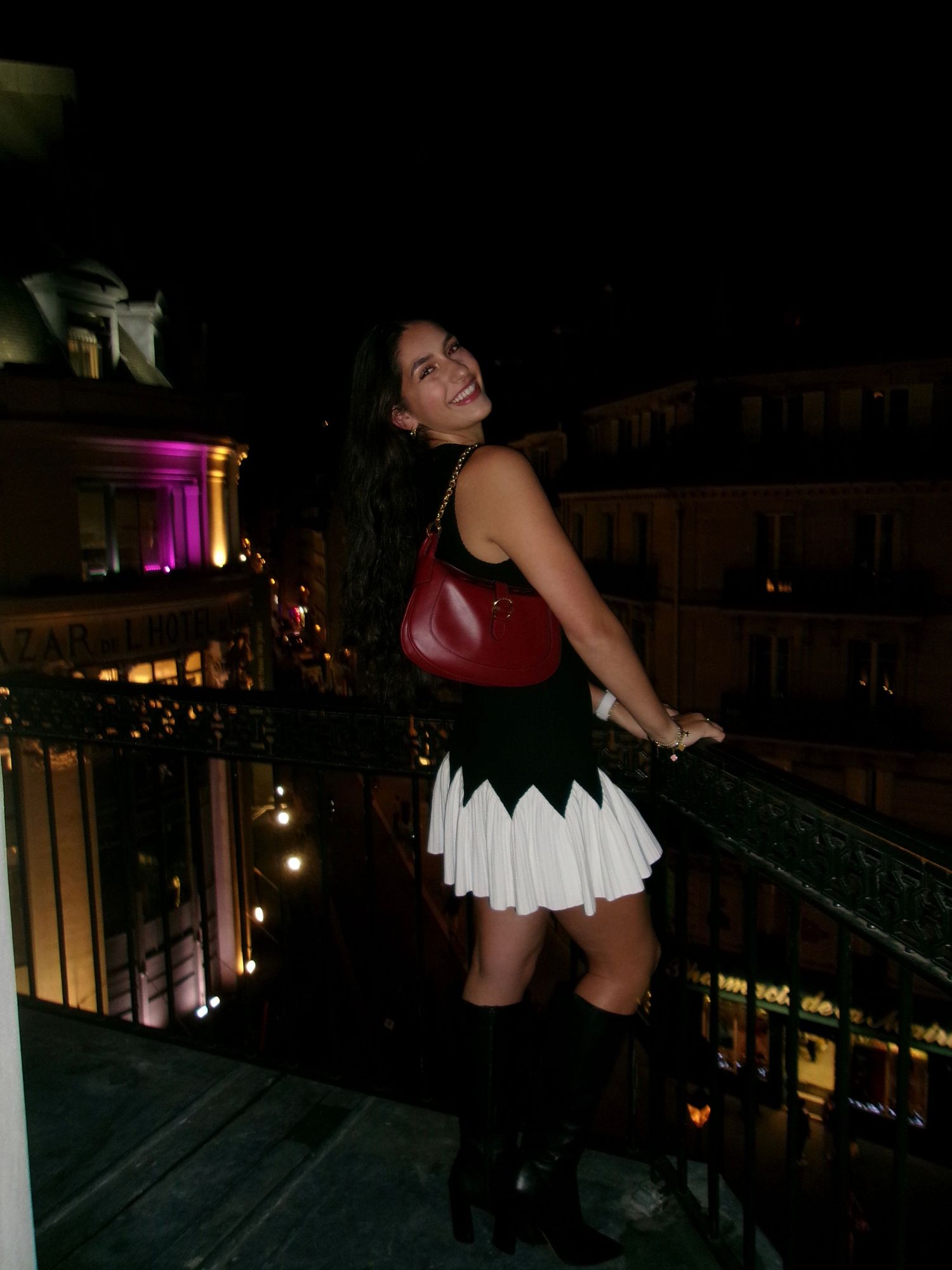 Woman in black and white skirt outfit smiles on a balcony overlooking illuminated city streets at night.