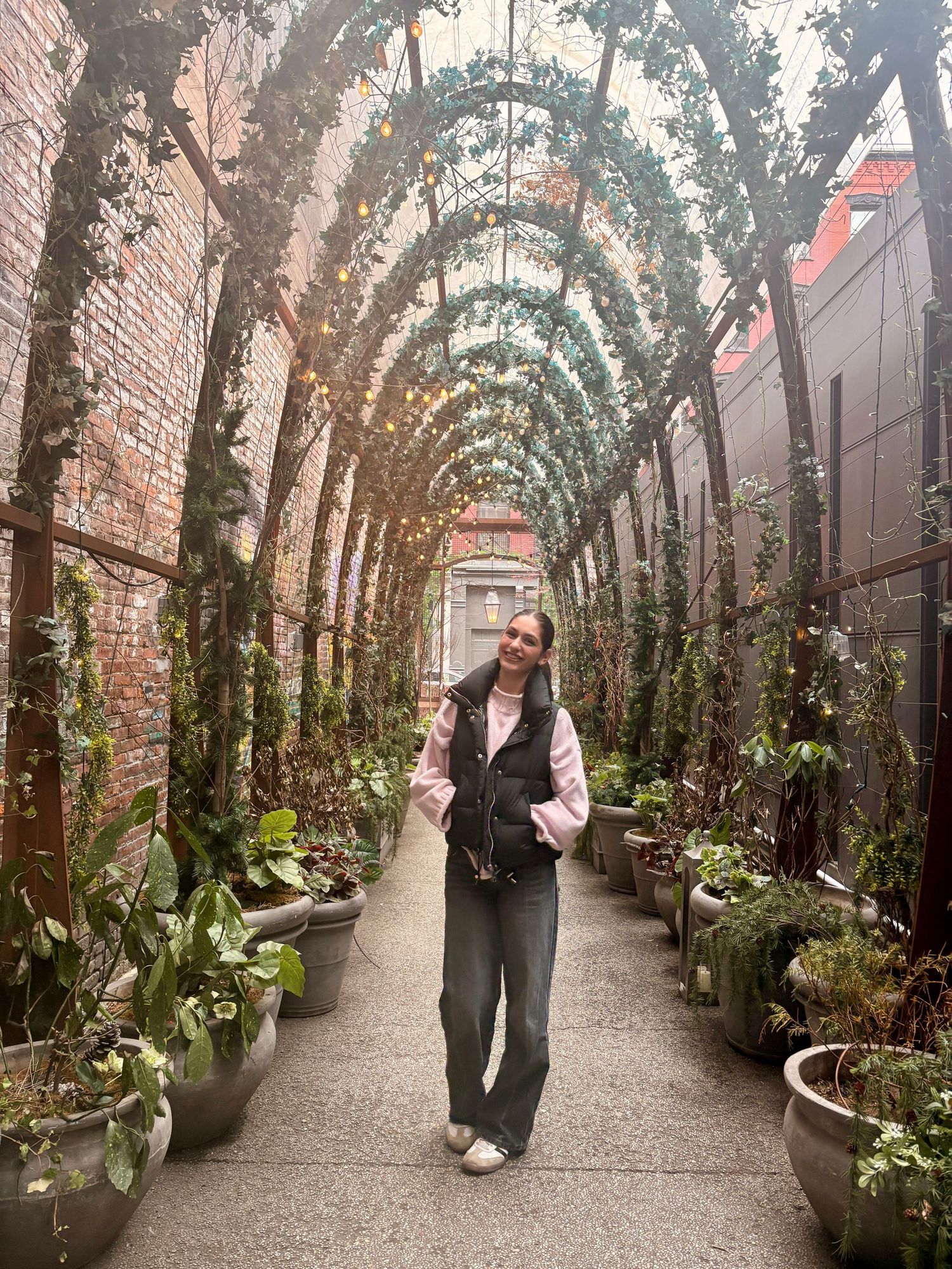Woman walking through arched garden tunnel with lush plants and string lights overhead.