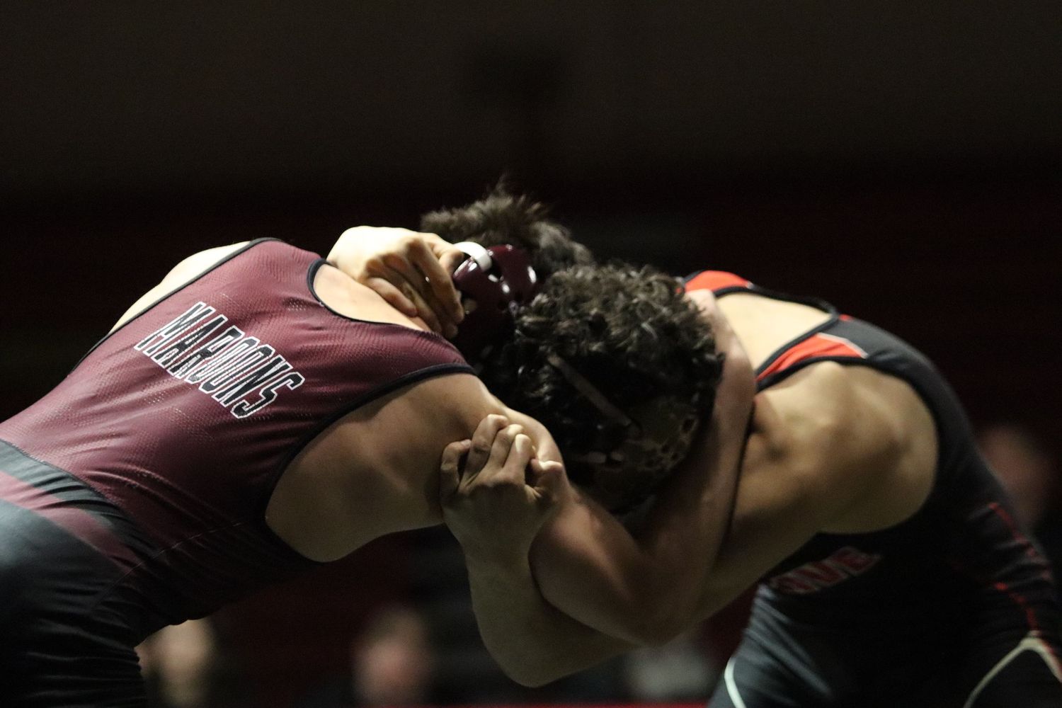 Two wrestlers grapple intensely during a high school wrestling match, wearing maroon and black uniforms.