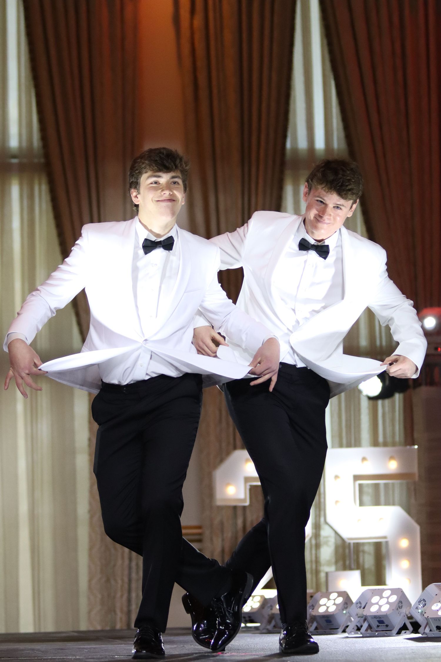 Two young men in matching white tuxedo jackets perform a synchronized dance routine on stage.
