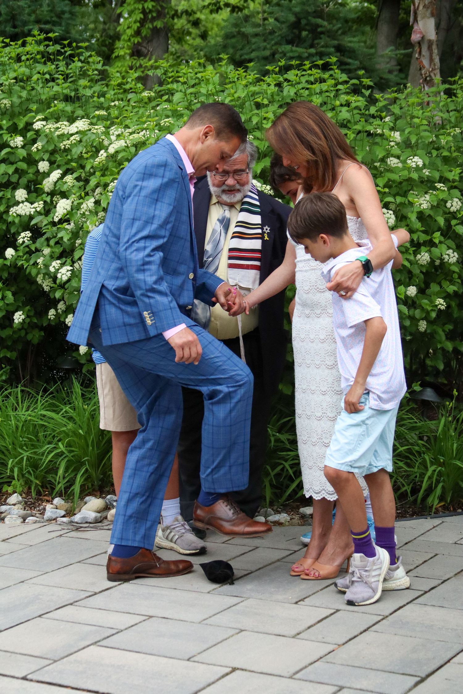 Groom in blue suit stomping glass during outdoor Jewish wedding ceremony with family.