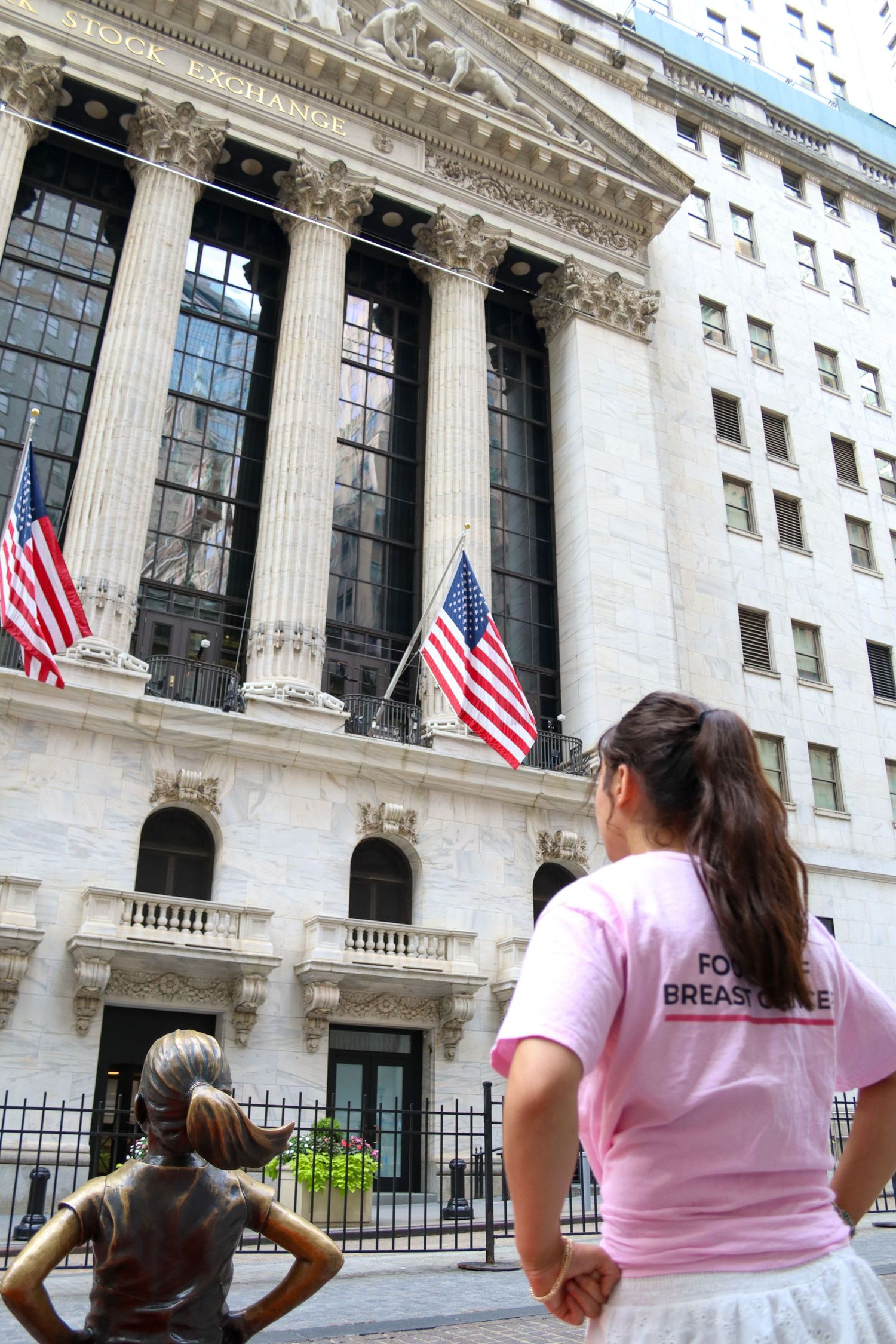 Woman in pink breast cancer shirt views Fearless Girl statue at NYSE Wall Street.