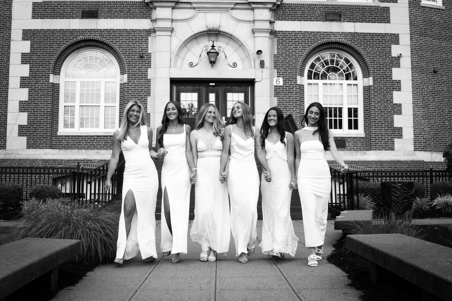 Group of seven women in white formal gowns posing in front of brick building.