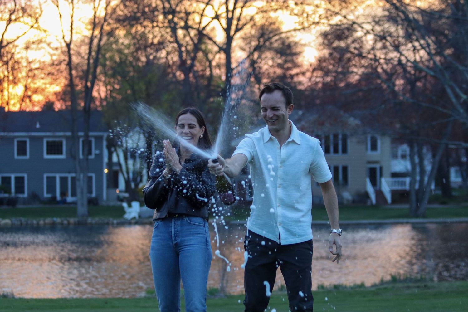 Couple celebrating at sunset by lake, popping champagne with water splashing dramatically.