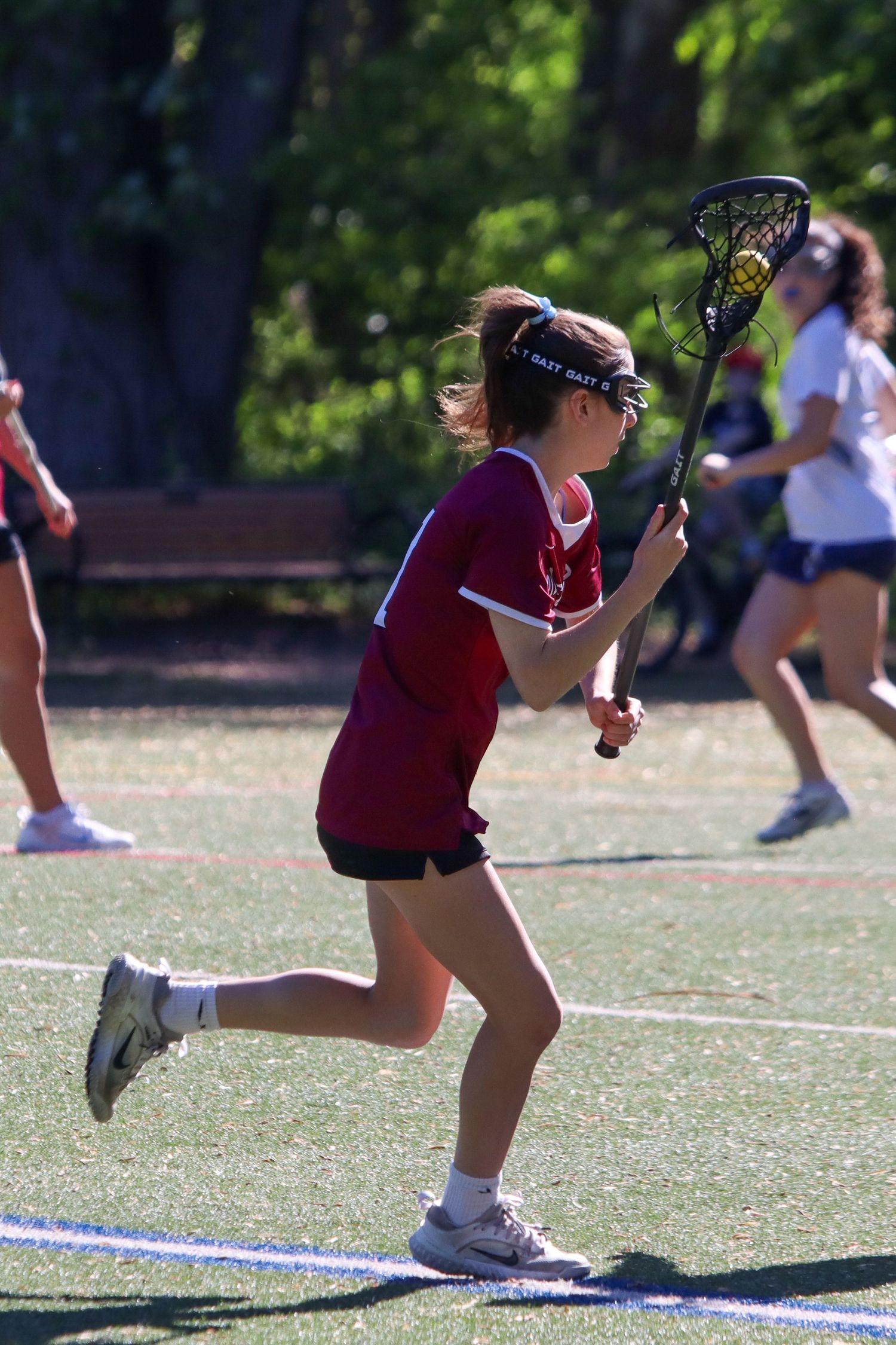 Young female lacrosse player in maroon uniform running with stick during outdoor game.
