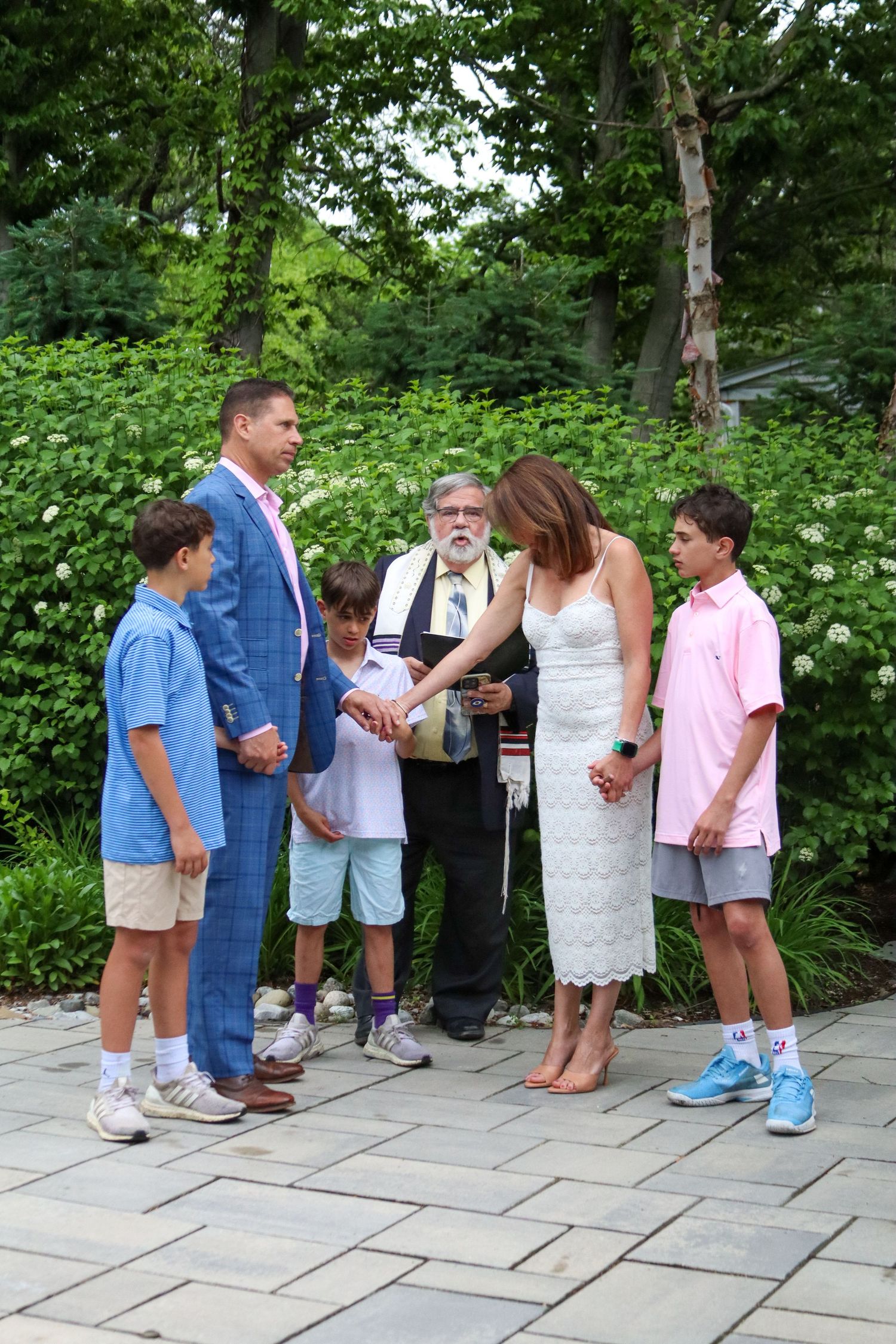 Family gathering outdoors for a Jewish wedding ceremony with officiant in lush garden setting.
