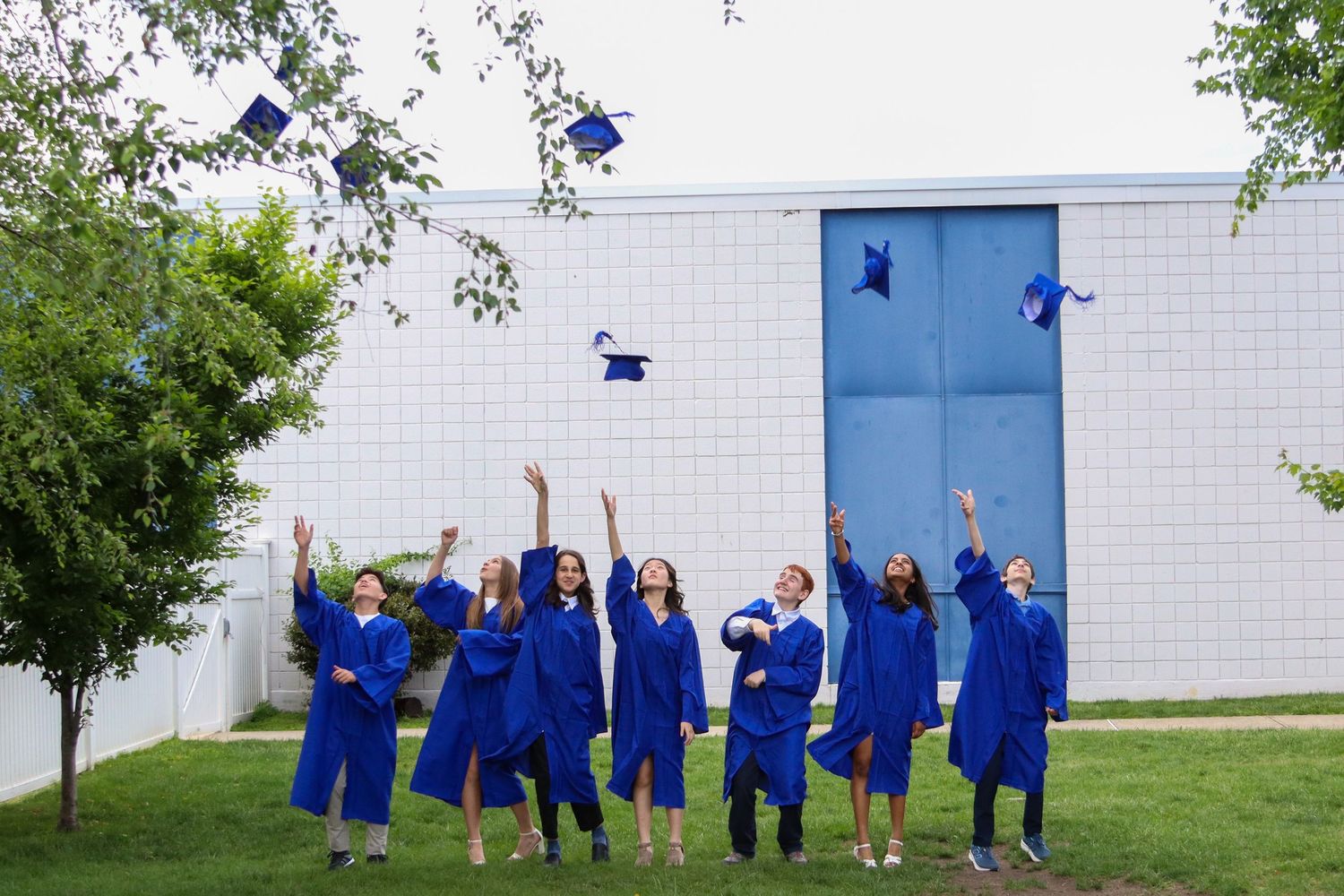 Group of graduates in blue caps and gowns tossing caps in air outdoors.