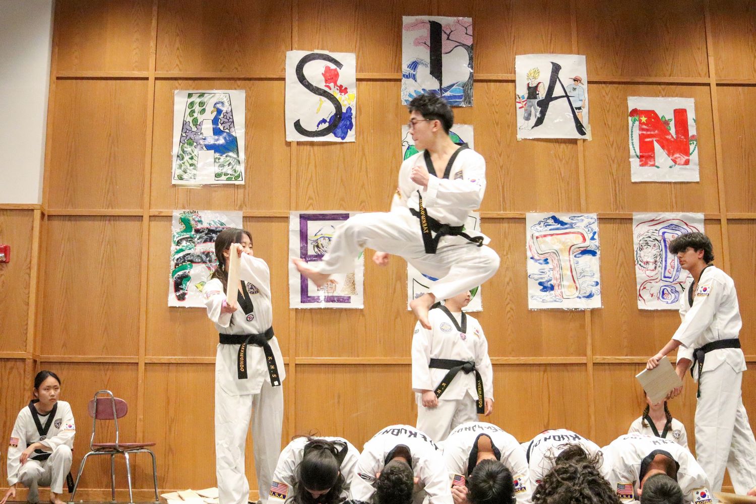 Taekwondo athlete performs a high jump kick over crouching students during a demonstration.