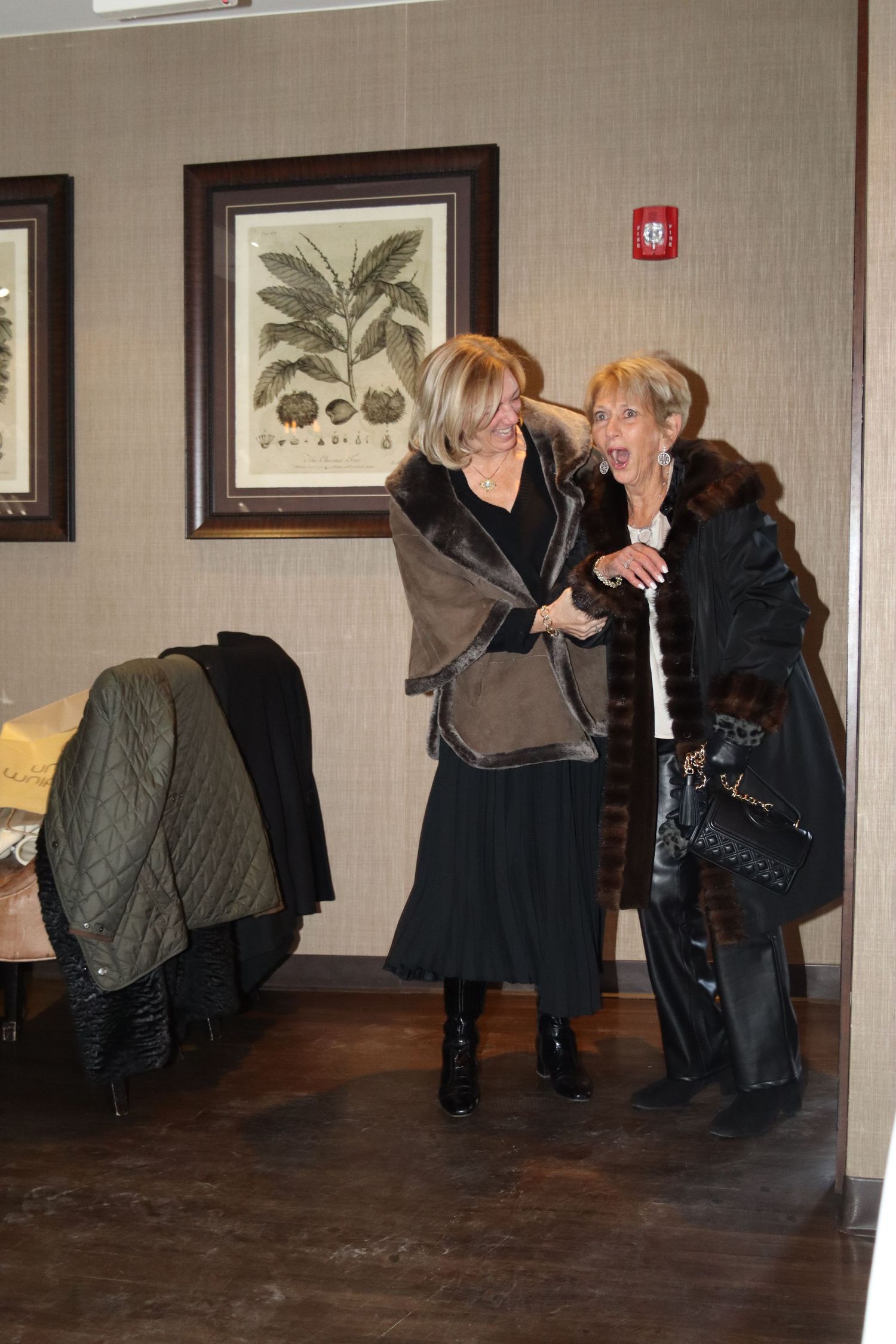 Two elegantly dressed older women in fur coats share a warm embrace indoors.