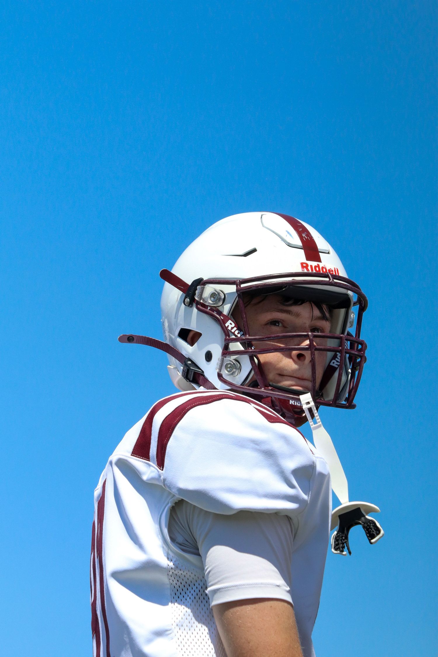 Young football player in white Riddell helmet and maroon uniform poses against a blue sky.