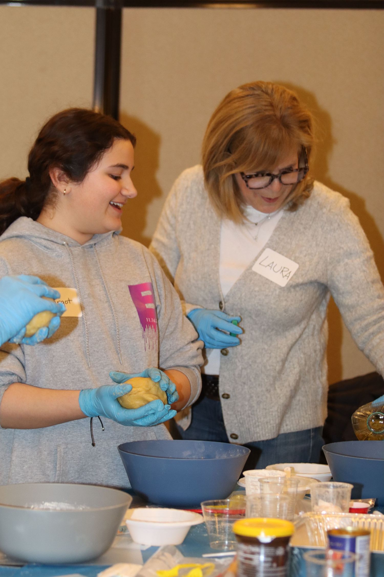 Two women wearing blue gloves preparing dough together at a cooking class event.