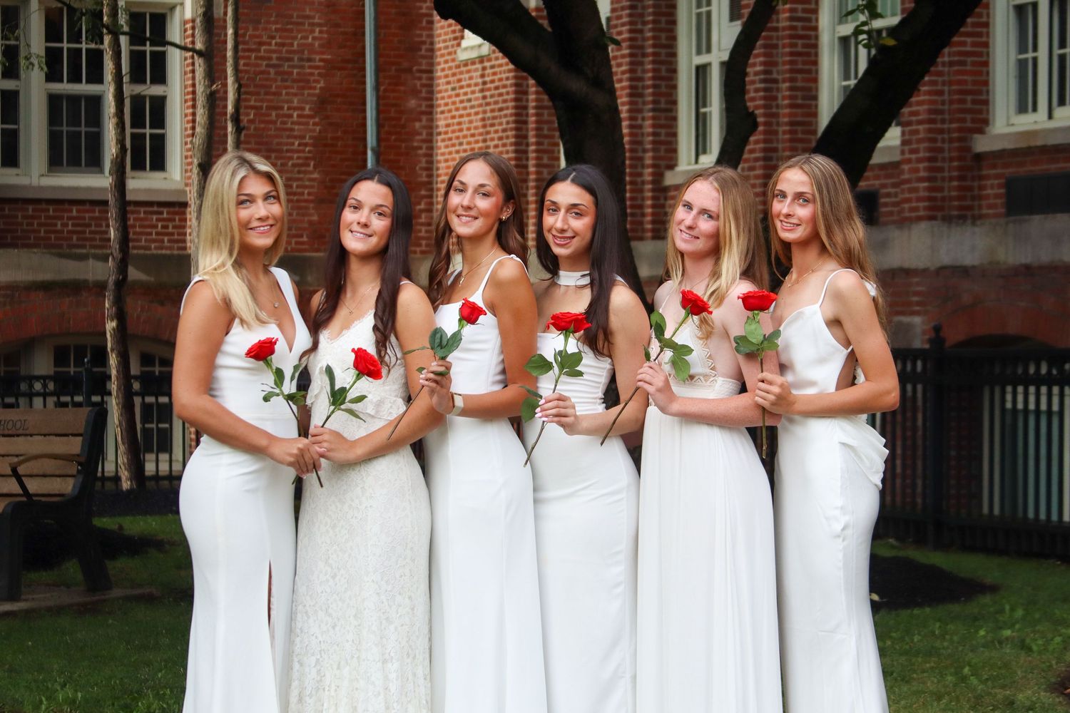 Six young women in white dresses holding red roses posing in front of brick building.