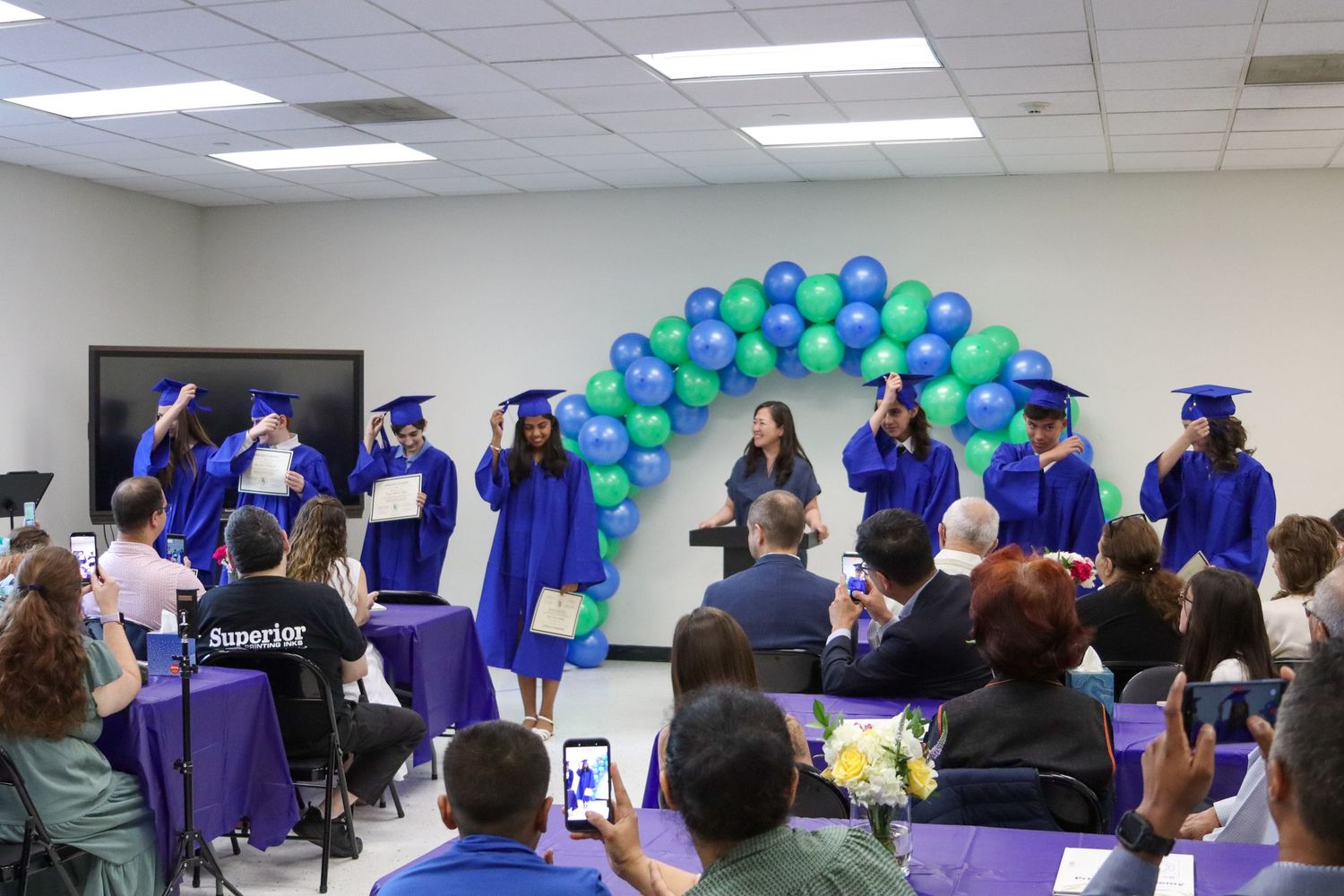 Graduates in blue caps and gowns celebrating under balloon arch at graduation ceremony.
