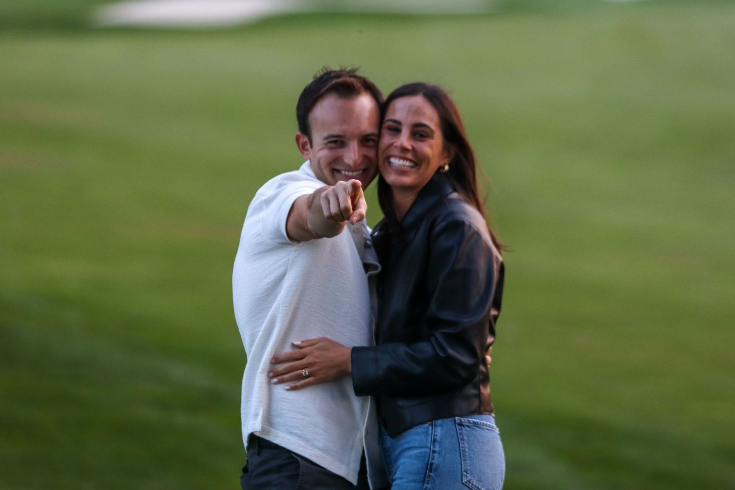 Happy couple embracing on golf course green, man pointing at camera, woman smiling.