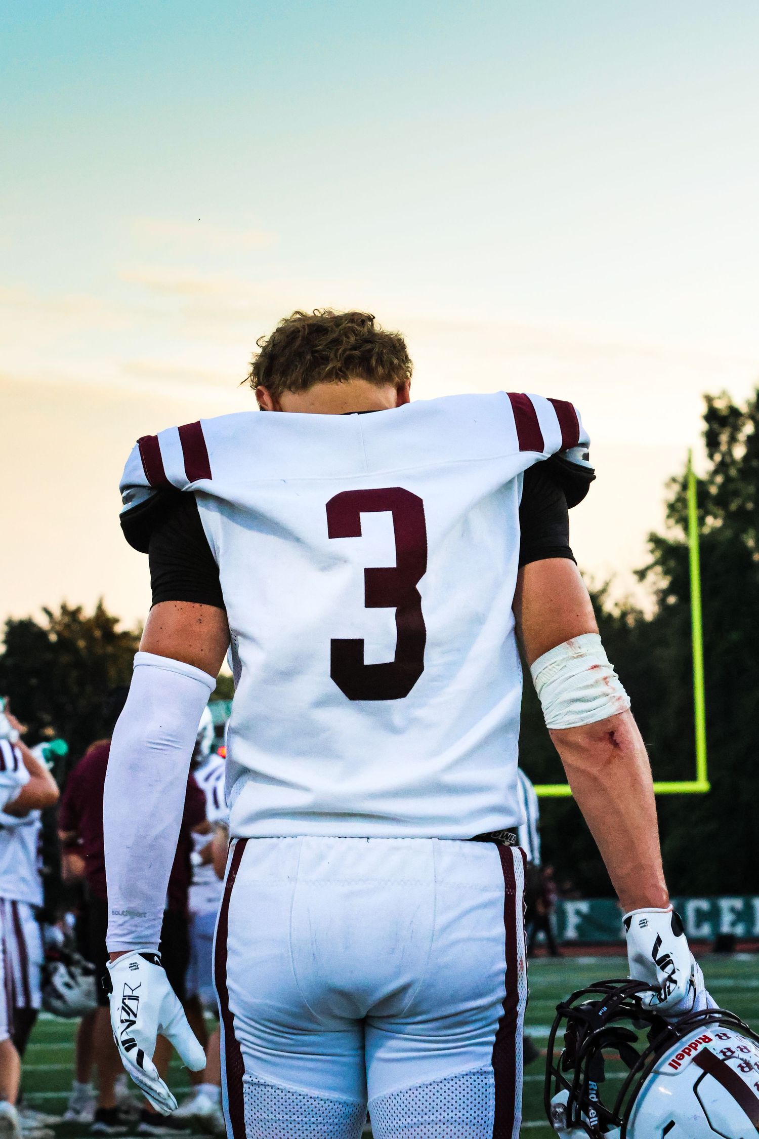 Football player wearing white jersey number 3 stands on field at sunset holding helmet.