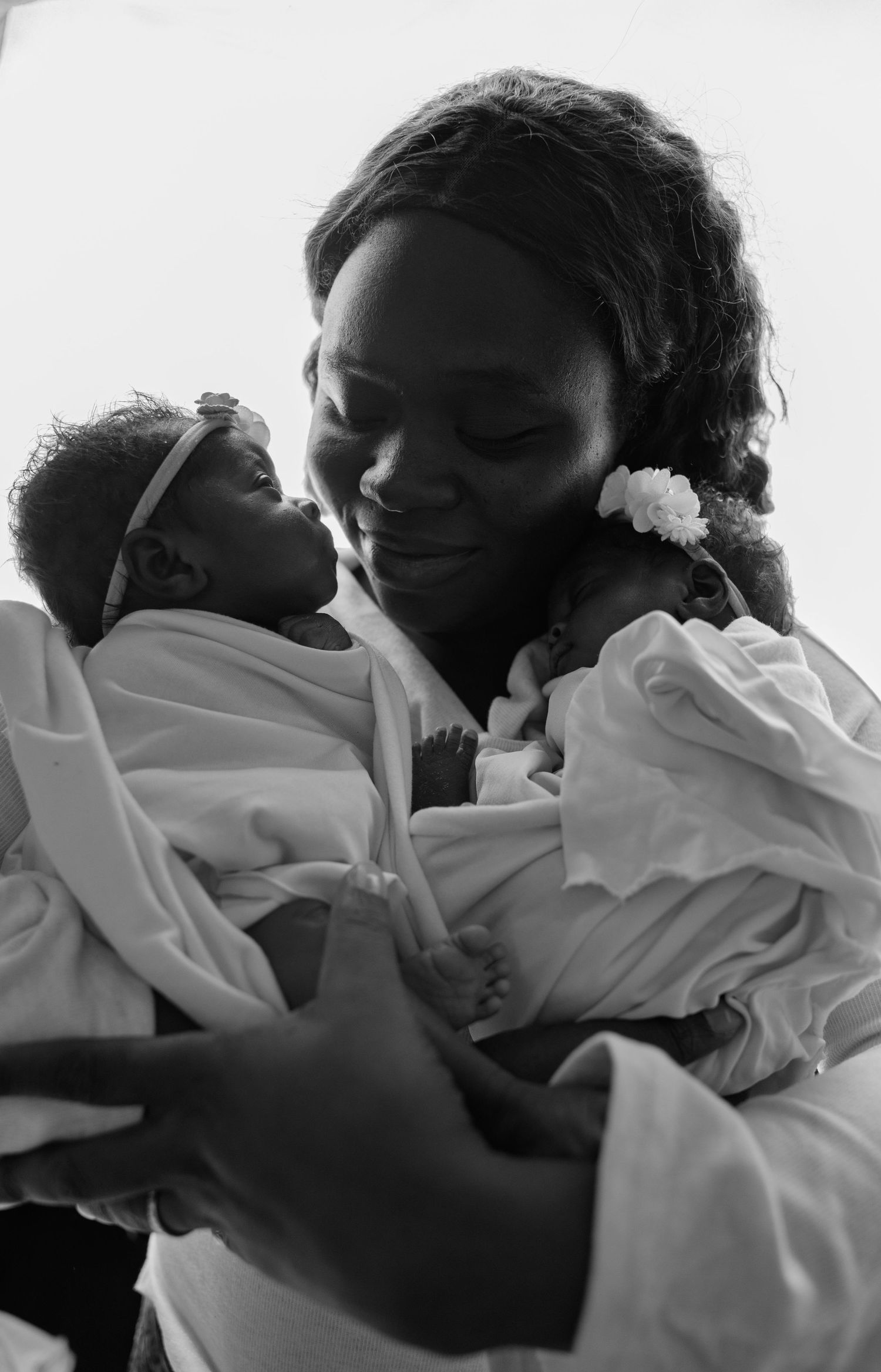 Black and white photo of a smiling mother tenderly holding two newborn twins wrapped in white blankets.