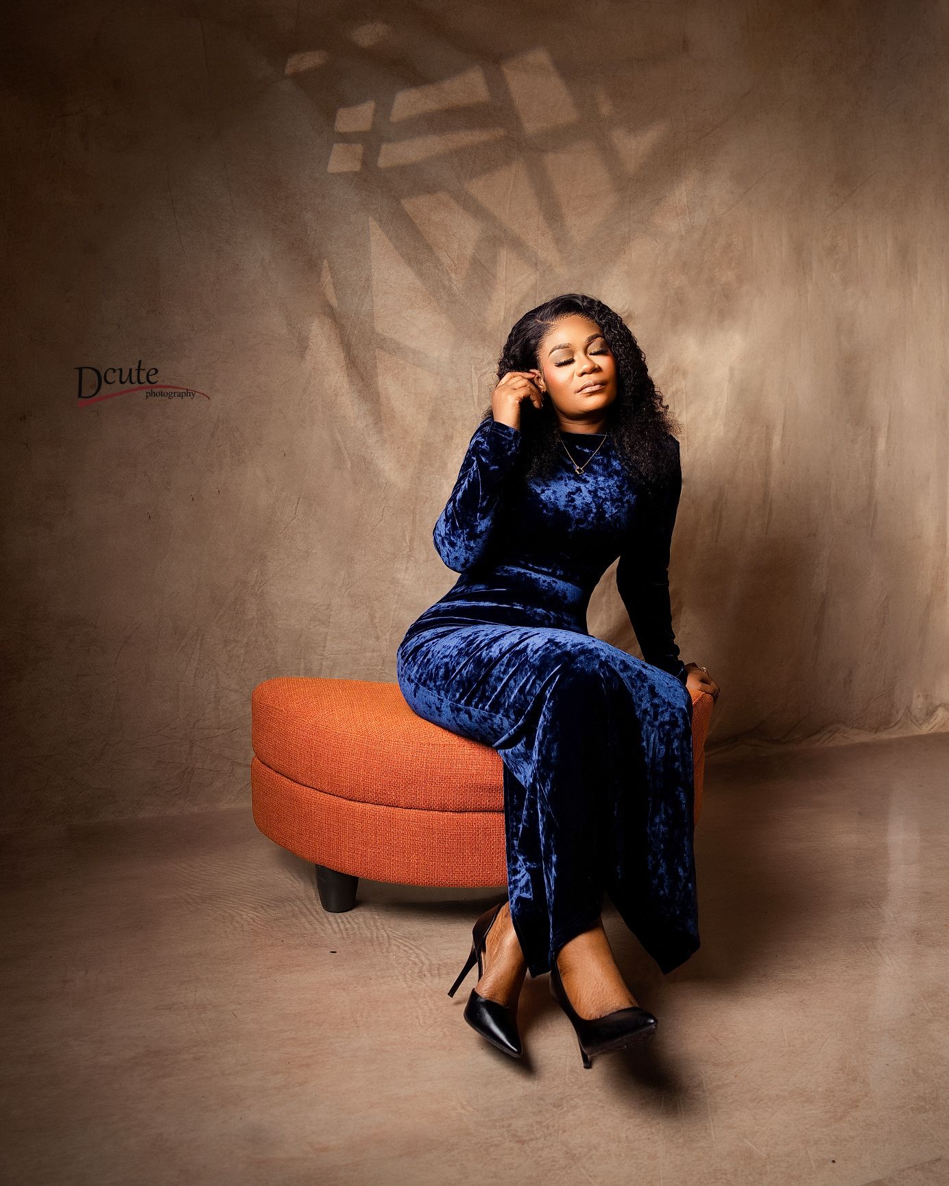 Woman in blue velvet dress seated on orange ottoman in moody studio portrait.