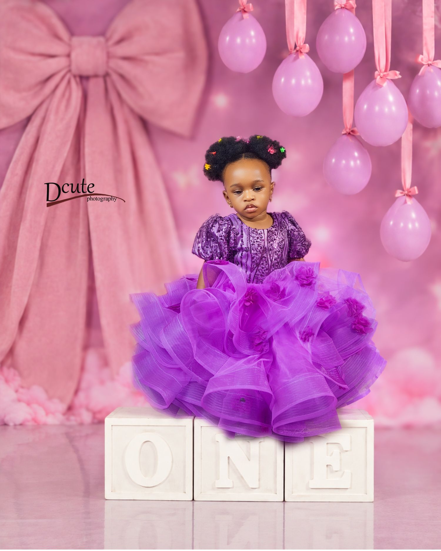Toddler girl in purple ruffled gown standing on ONE letter blocks with pink balloons and bow backdrop.