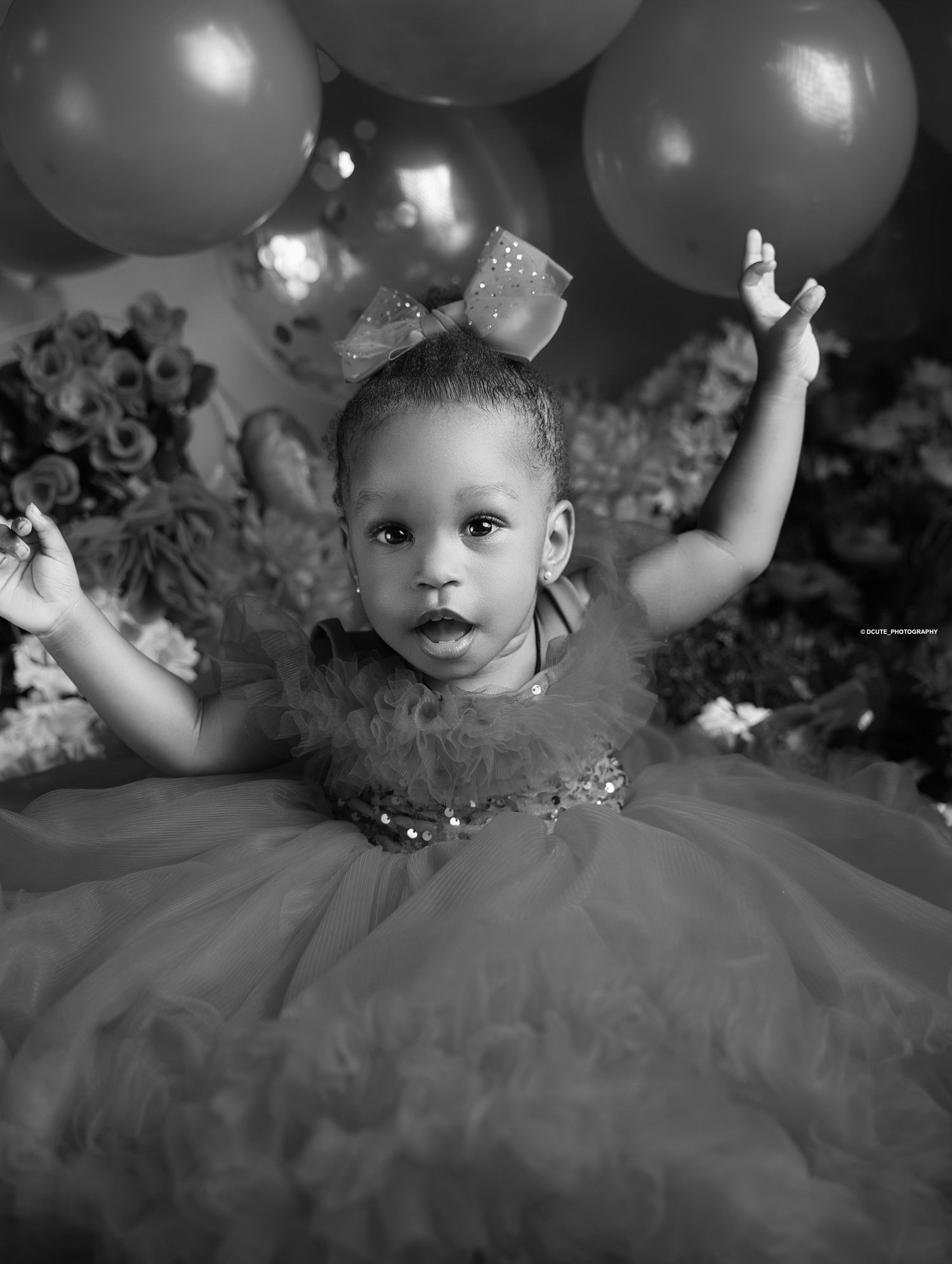 Black and white photo of baby girl in tutu dress with bow raising arms among balloons during toddler photoshoot in North York