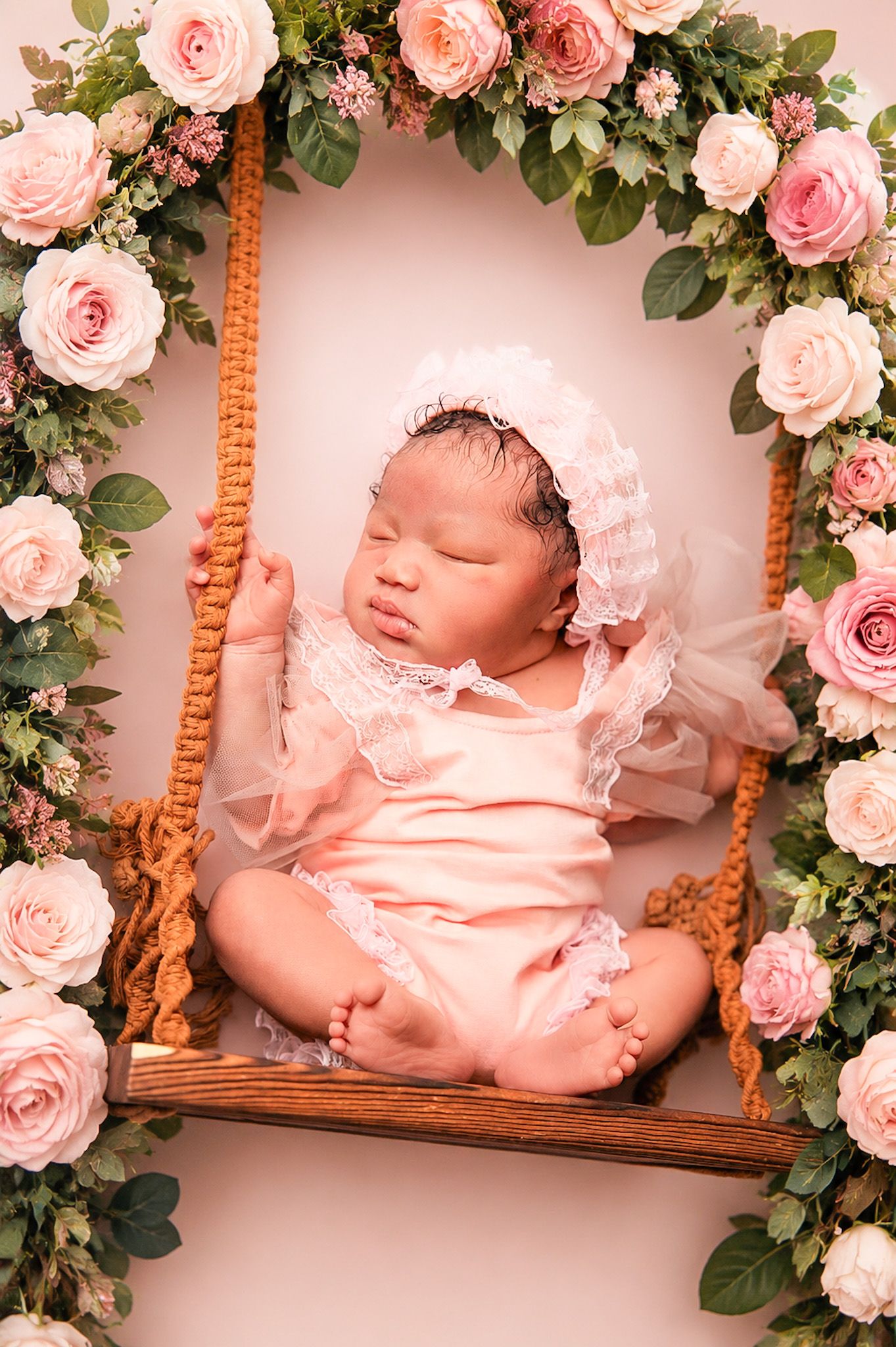 Newborn baby girl sleeping peacefully on a wooden swing surrounded by pink roses in a floral portrait.