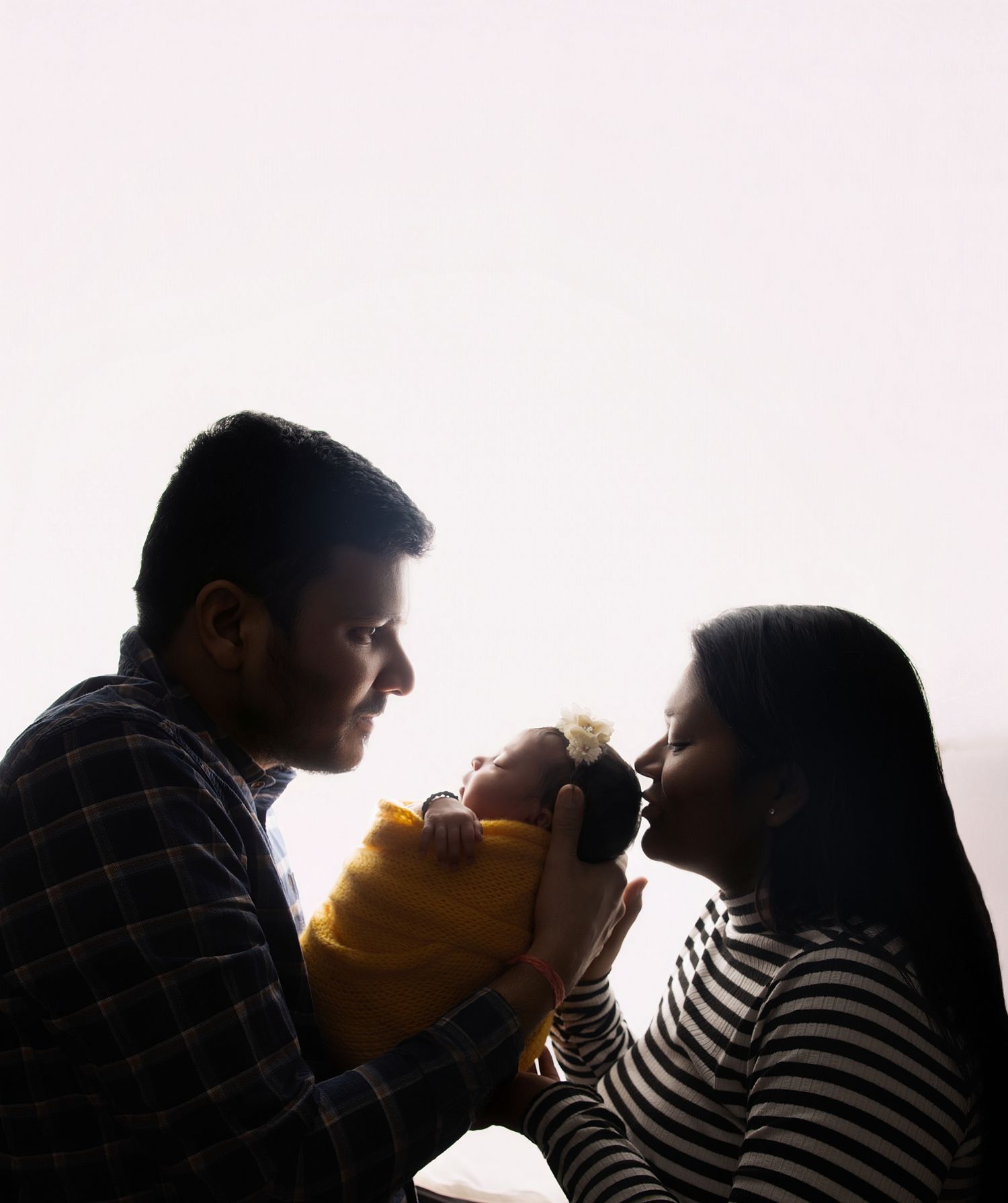 Silhouetted parents gaze lovingly at their newborn baby wrapped in yellow against a bright window.