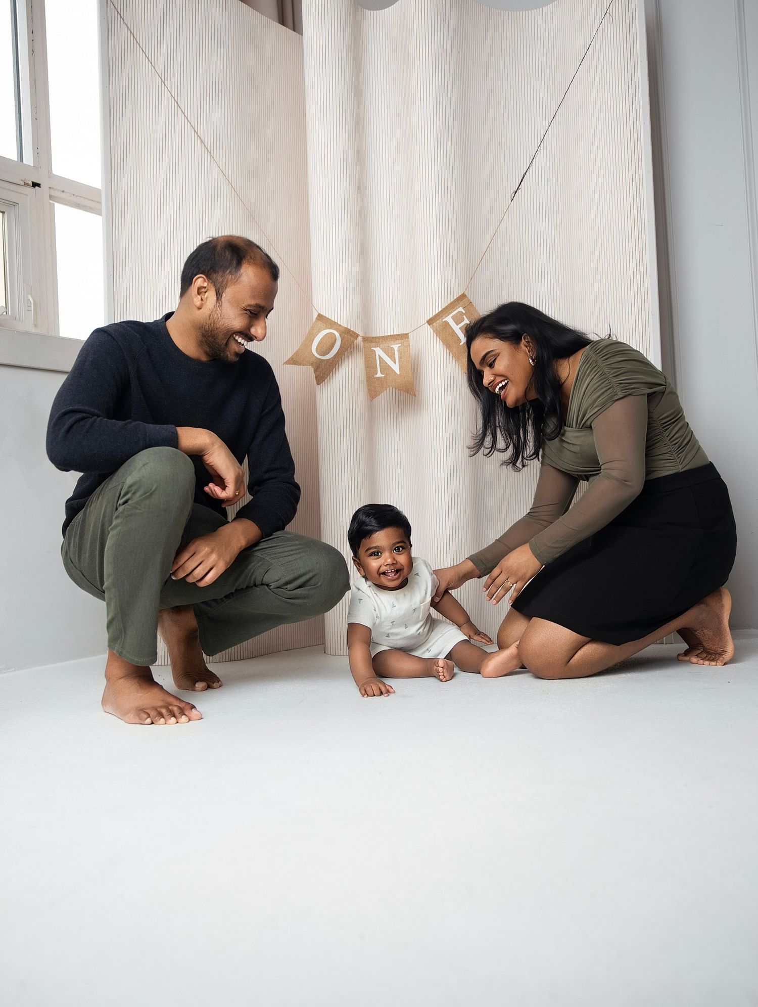 Indian parents crouching beside baby sitting on white floor under ONE banner during first birthday photo session.